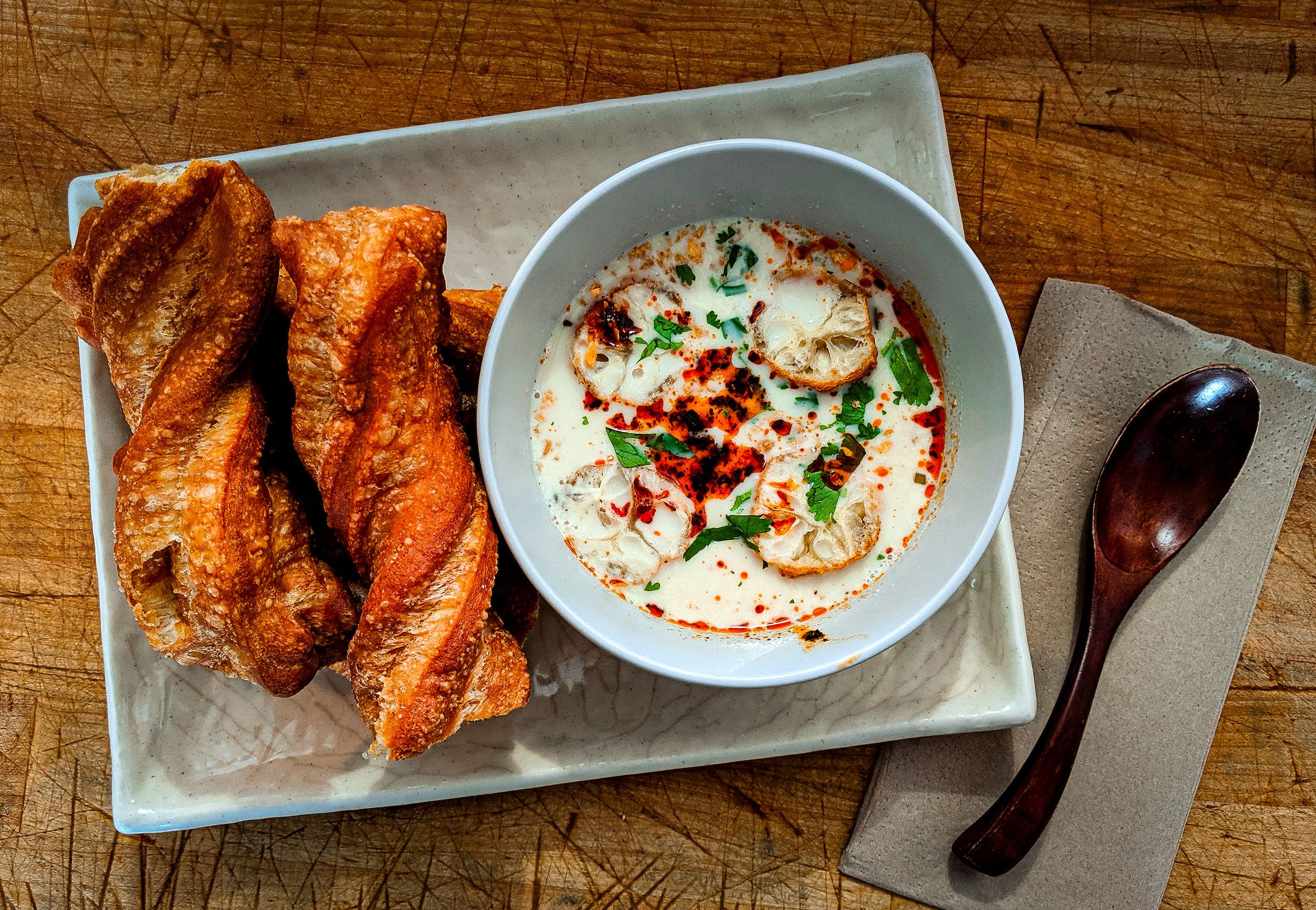 Crusty baguette slices beside a bowl of creamy soup loaded with herbs and croutons, drizzled with chili oil, on a wooden table; a spoon rests on a napkin nearby.