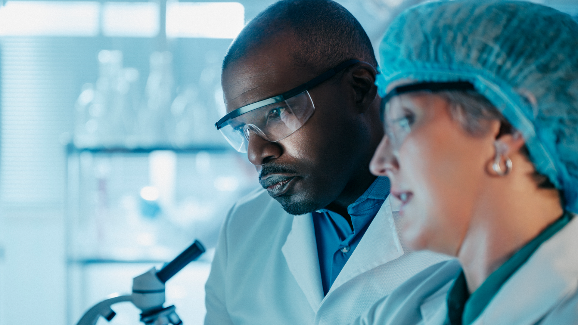Two scientists in a laboratory wearing white coats and safety goggles. A man studies at a microscope while a woman with a blue hairnet observes, under cool blue lighting.