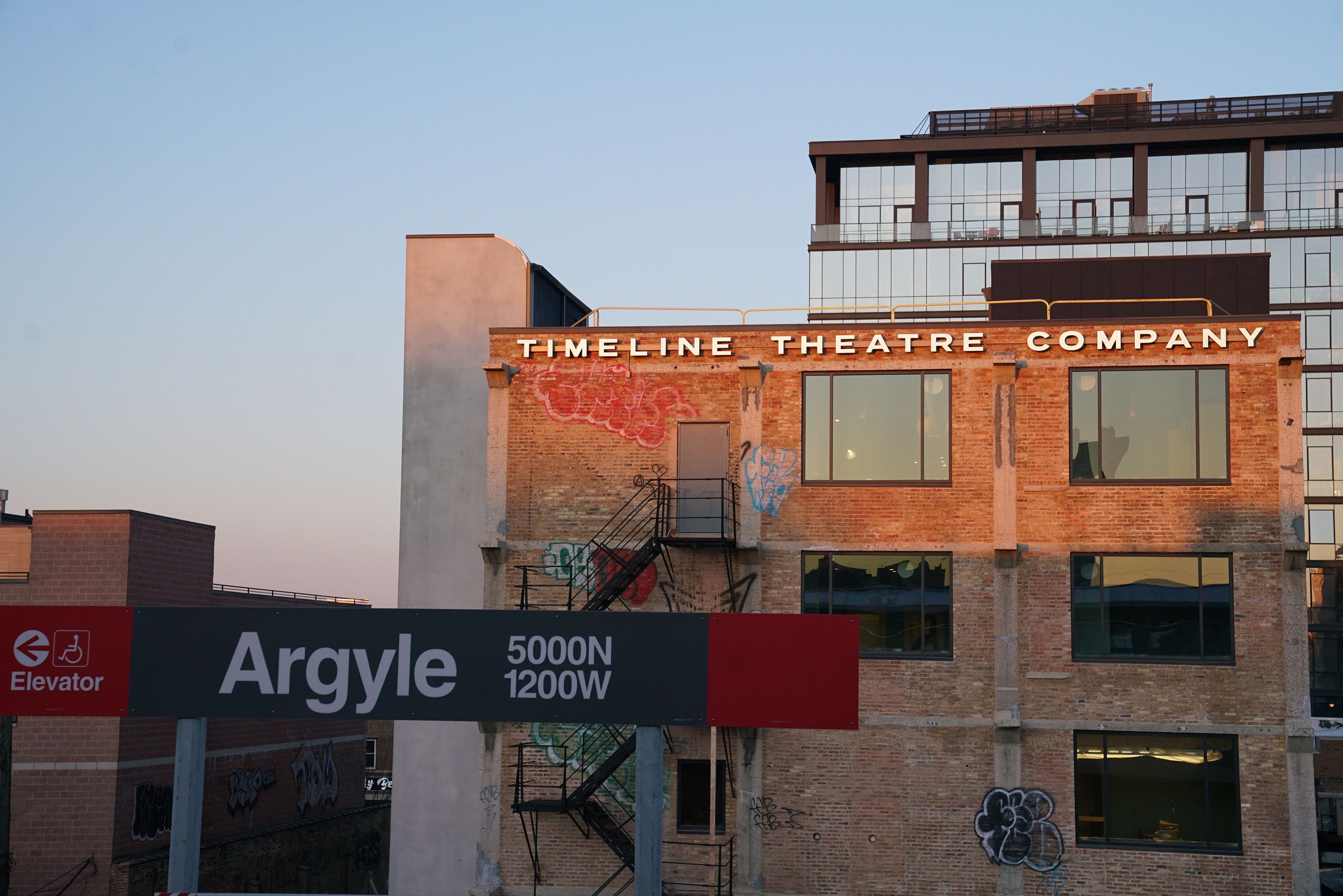 Brick building with a sign reading "Timeline Theatre Company" atop. In front, a large "Argyle" sign with 5000N 1200W and an elevator icon. Graffiti walls, a metal fire escape, and a glass high-rise behind at sunset.