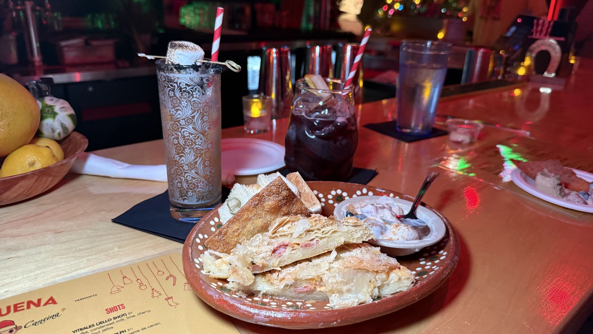 Plate with fried snacks and dipping sauce on a bar counter with two drinks, one with a striped straw and toasted marshmallow garnish. Christmas lights and tree in background.