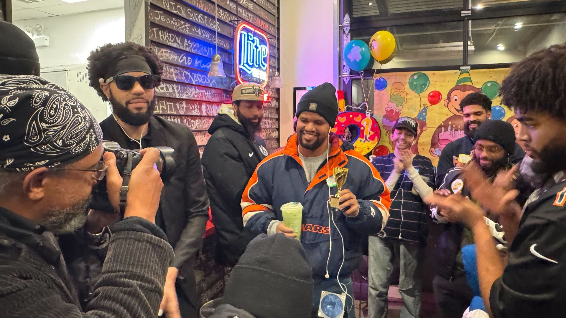 Group of people indoors celebrating; man in orange and navy jacket holding trophy and green drink; others clapping and smiling; colorful balloons and party decorations in background.