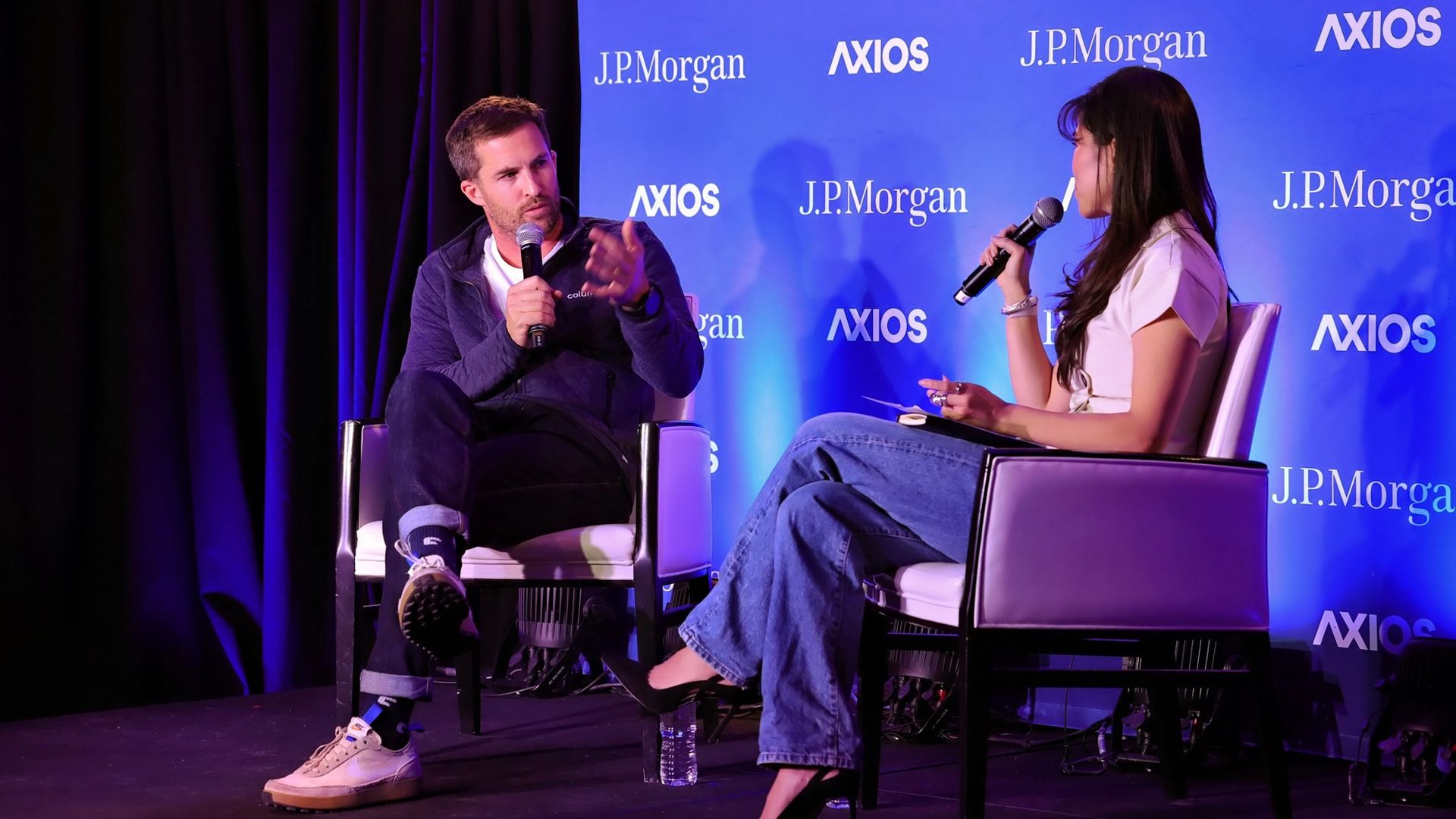 Two people seated on stage in a discussion, holding microphones. Blue backdrop with J.P. Morgan and AXIOS logos. Man wears a dark jacket, woman in white top and jeans.