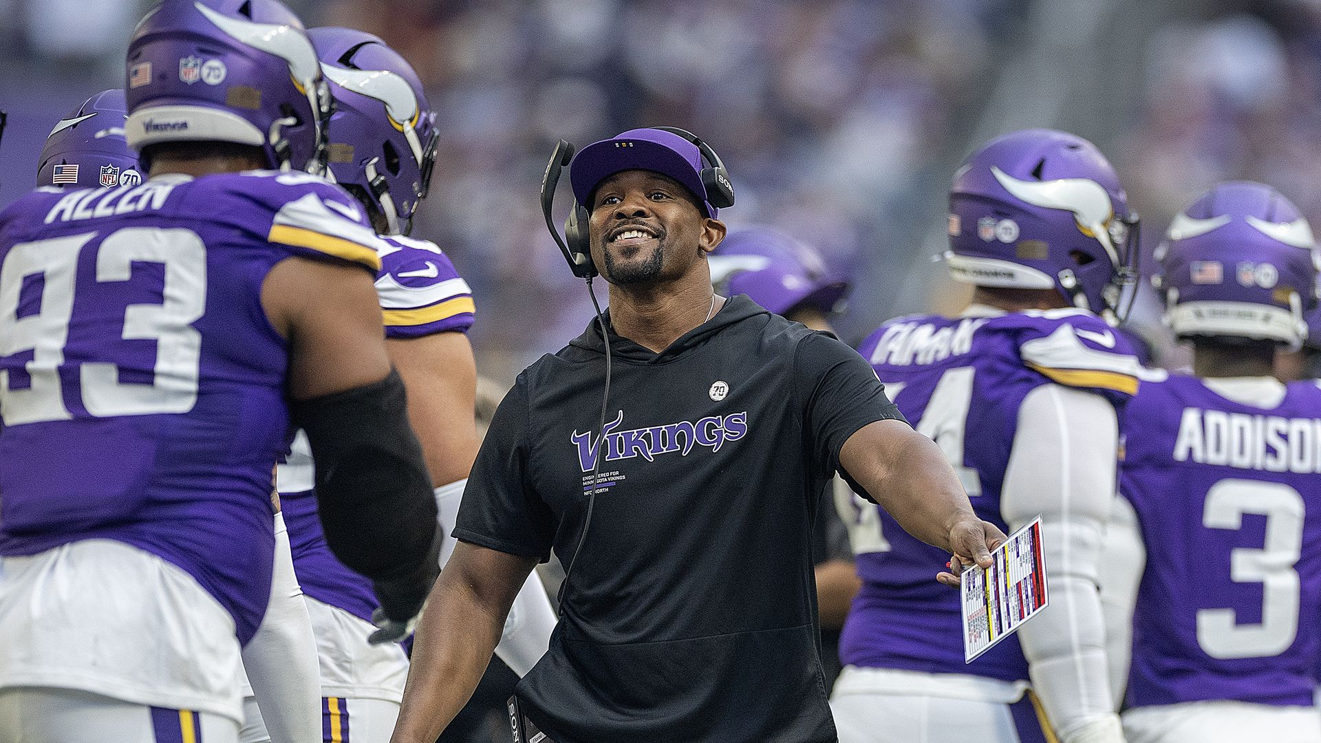 Brian Flores, wearing a black Vikings shirt and purple hat, smiles and celebrates with players 