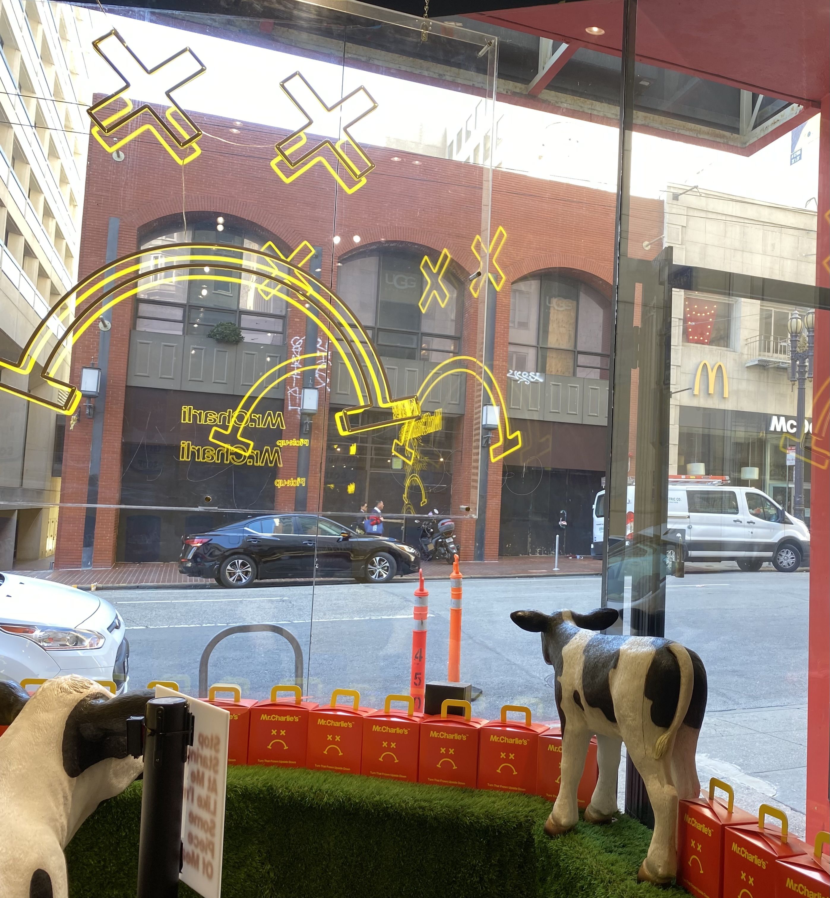A photo showing yellow illuminated lights in the shape of a frowning face on a window. Below the lights are fake cows looking out the window. Across the street is a McDonald's logo.
