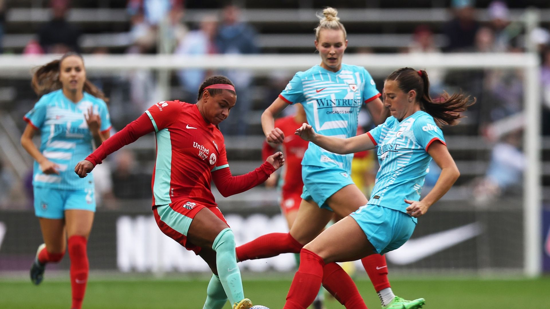 Four soccer players, three in blue uniforms, one in red uniform vying for a soccer ball.