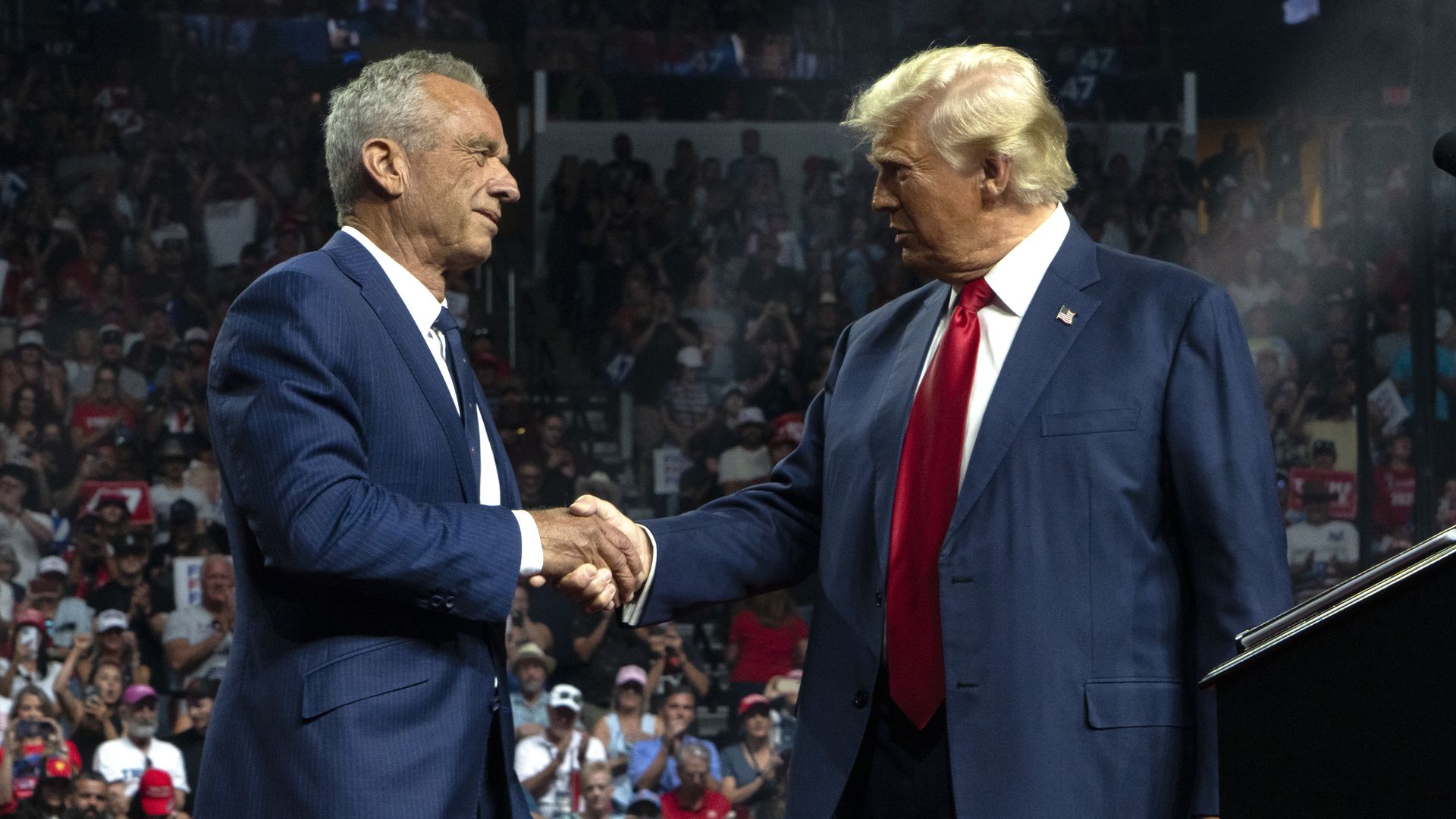 Trump and RFK Jr. shake hands in front of a rally crowd.