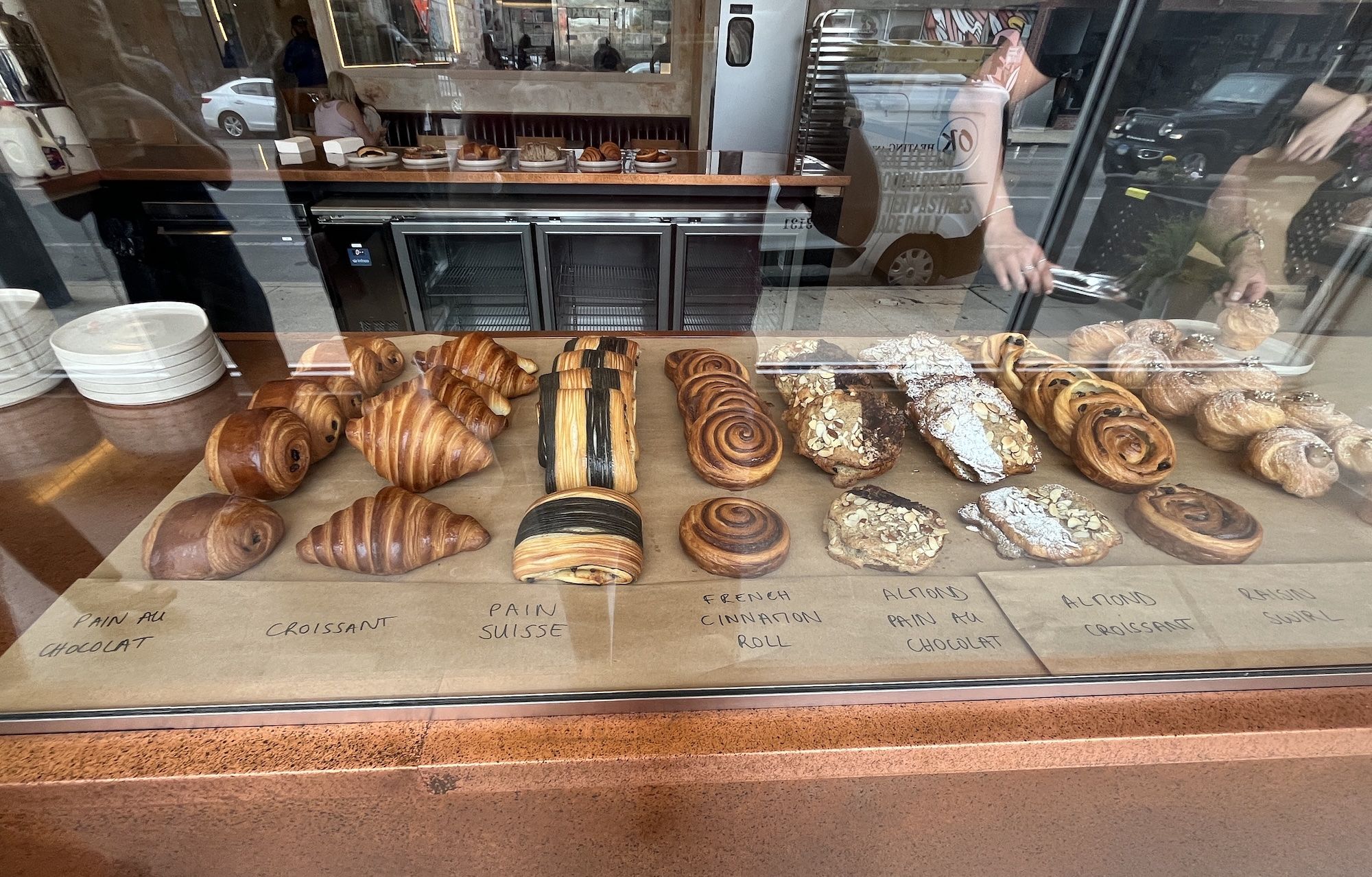 Bakery display case with assorted pastries—croissants, pains au chocolat, cinnamon rolls, almond croissants; handwritten labels on the counter; glass reflects street and passing cars outside.
