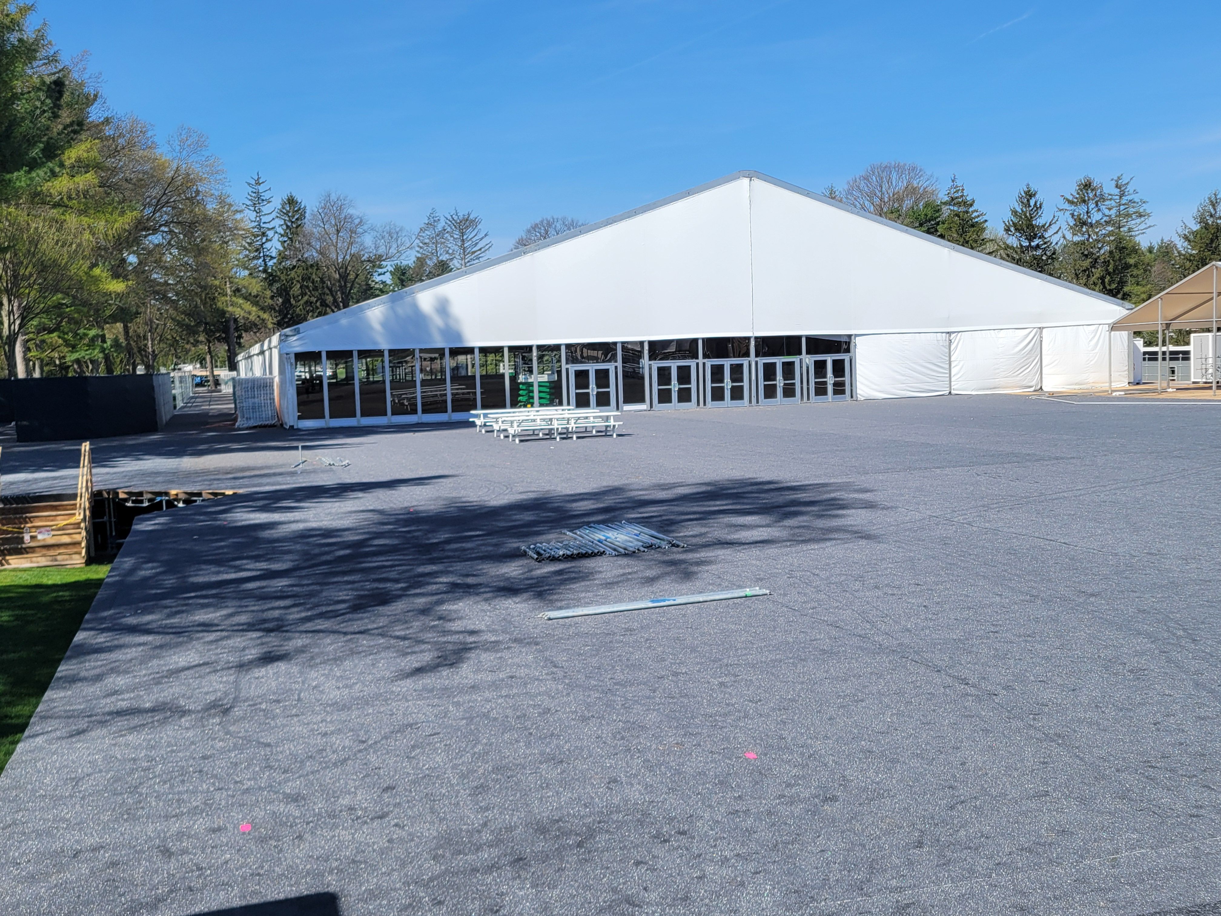 Large white event tent set up on a wide paved lot, with glass entrance doors, a few white picnic tables, and trees in the background under a clear blue sky.