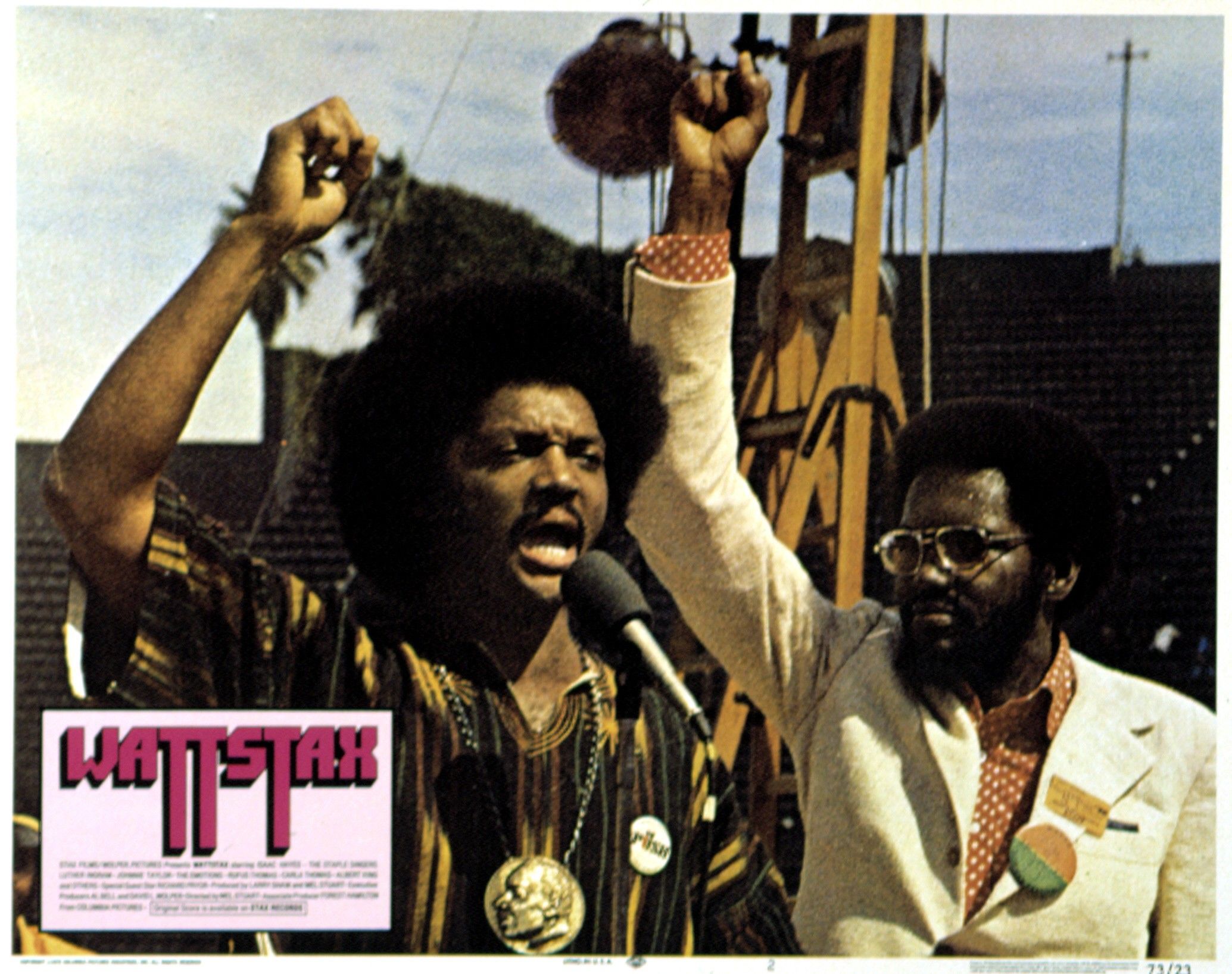 Two African American men raising fists, one speaking into a microphone, the other wearing sunglasses and a white suit with a button. Text reads "WATTSTAX" in pink and black.