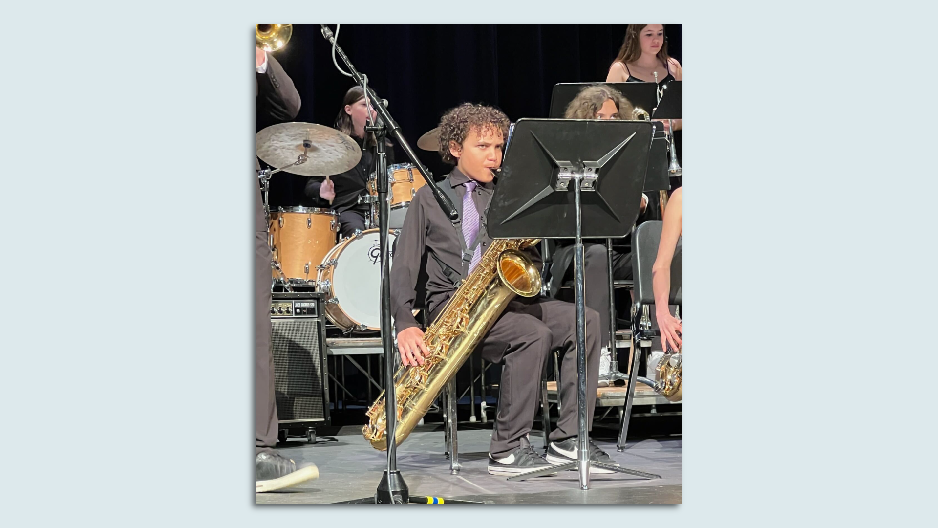 A young person in black pants and black button up shirt sits playing a saxophone with other band members nearby.