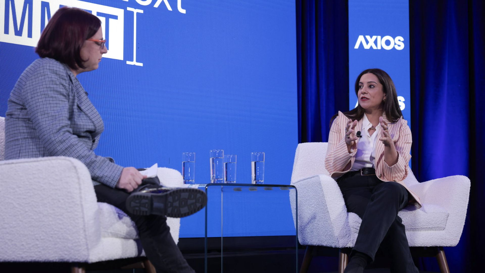 Ina Fried (left) wearing a gray jacket sits across from Reshma Saujani (right) wearing a striped pink jacket speaking with hands outstretched. Behind them is a blue backdrop with AXIOS written in white. 