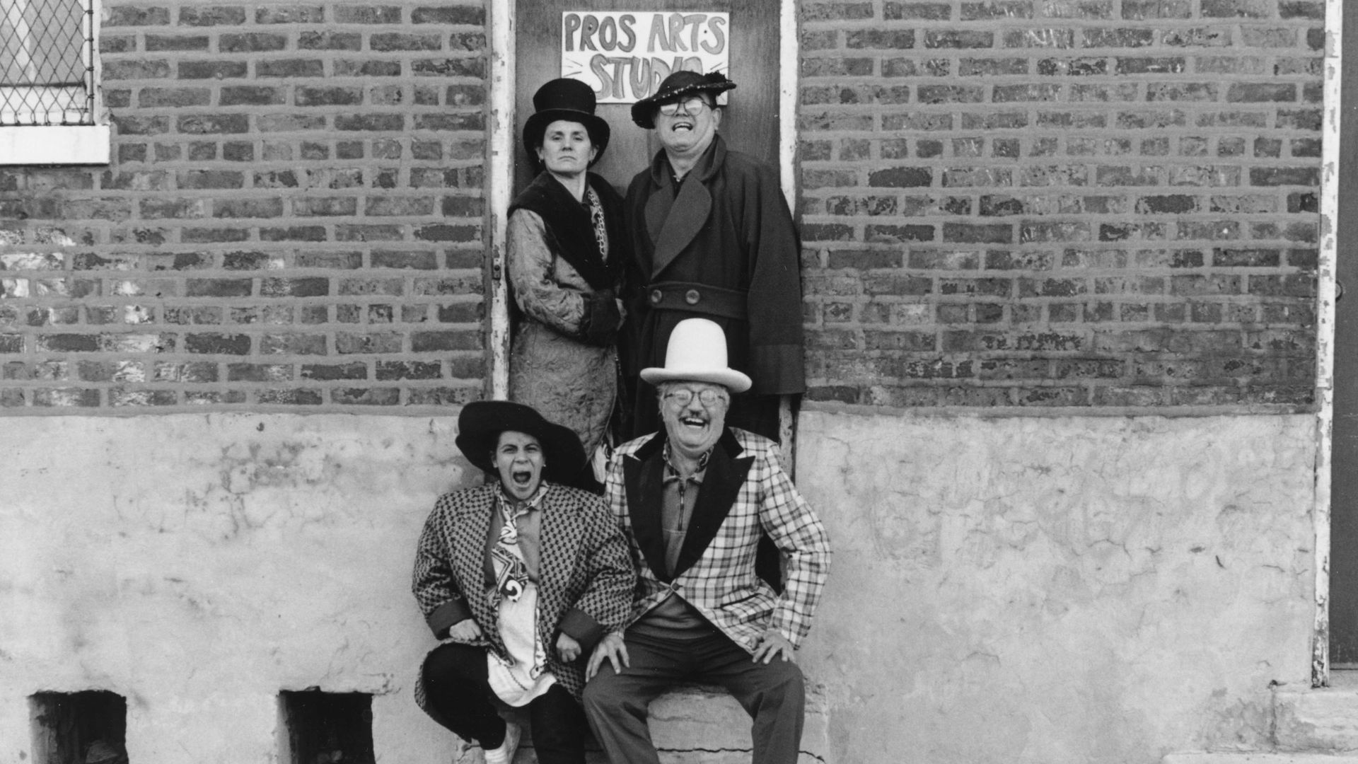 Black and white photo of four people wearing vintage hats and coats posing expressively in front of a brick wall and door labeled "Pros Arts Studio." Two sit on a step, two stand behind.