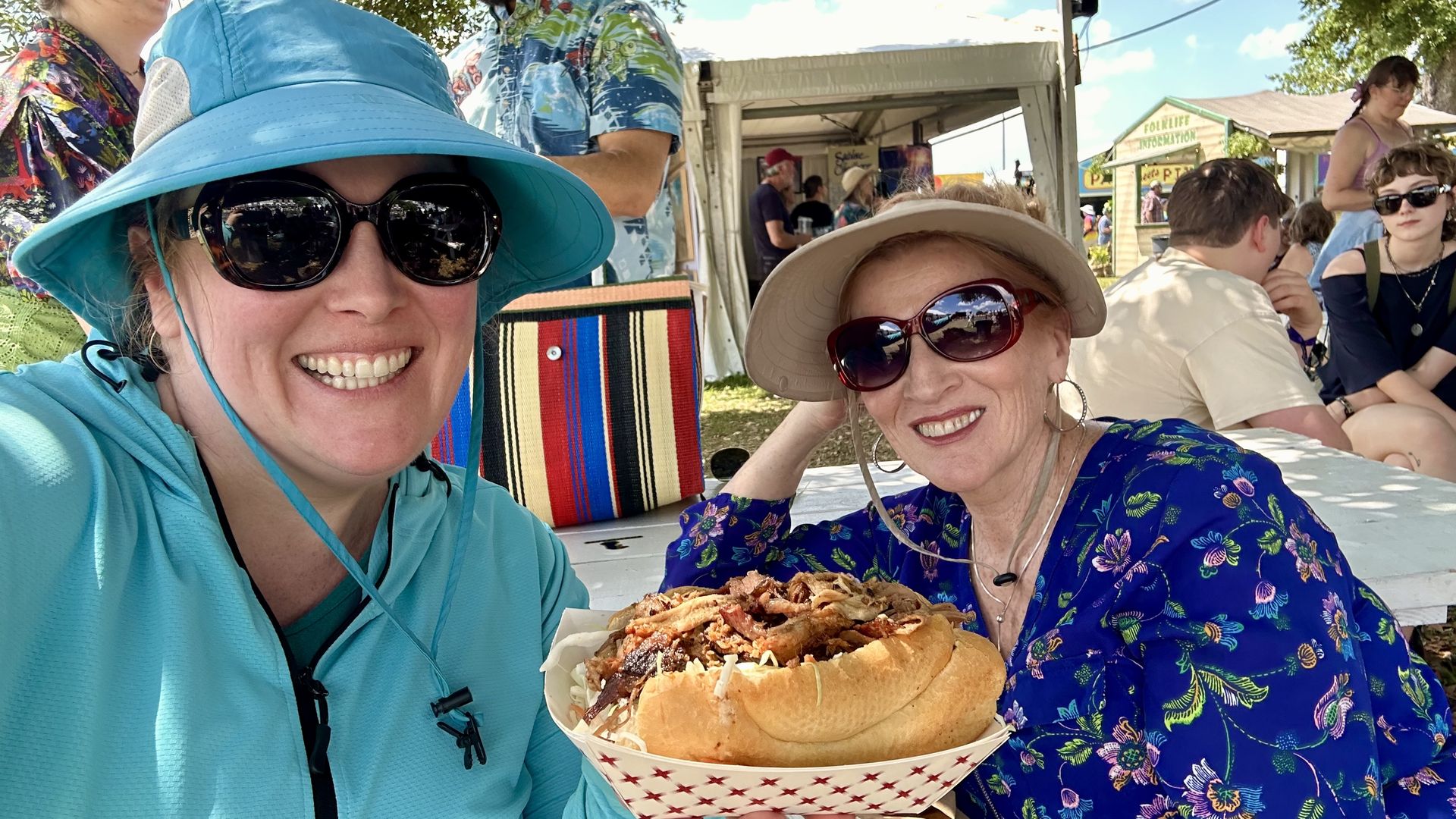 Two women in sunglasses and wide-brim hats smile at an outdoor festival, sharing a large sandwich. Left in turquoise jacket, right in beige hat and purple floral top, tents and people behind.