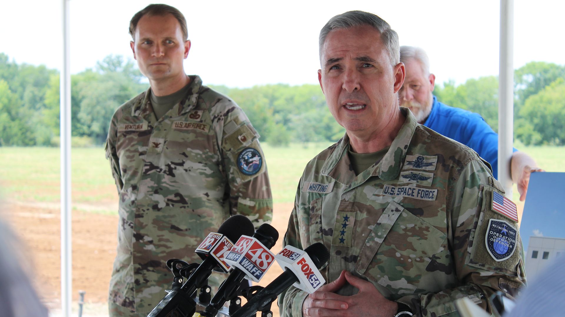 Three military officers under a tent at a press briefing. Front-right, a gray-haired Space Force officer speaks into microphones; left, another soldier stands; trees in background.