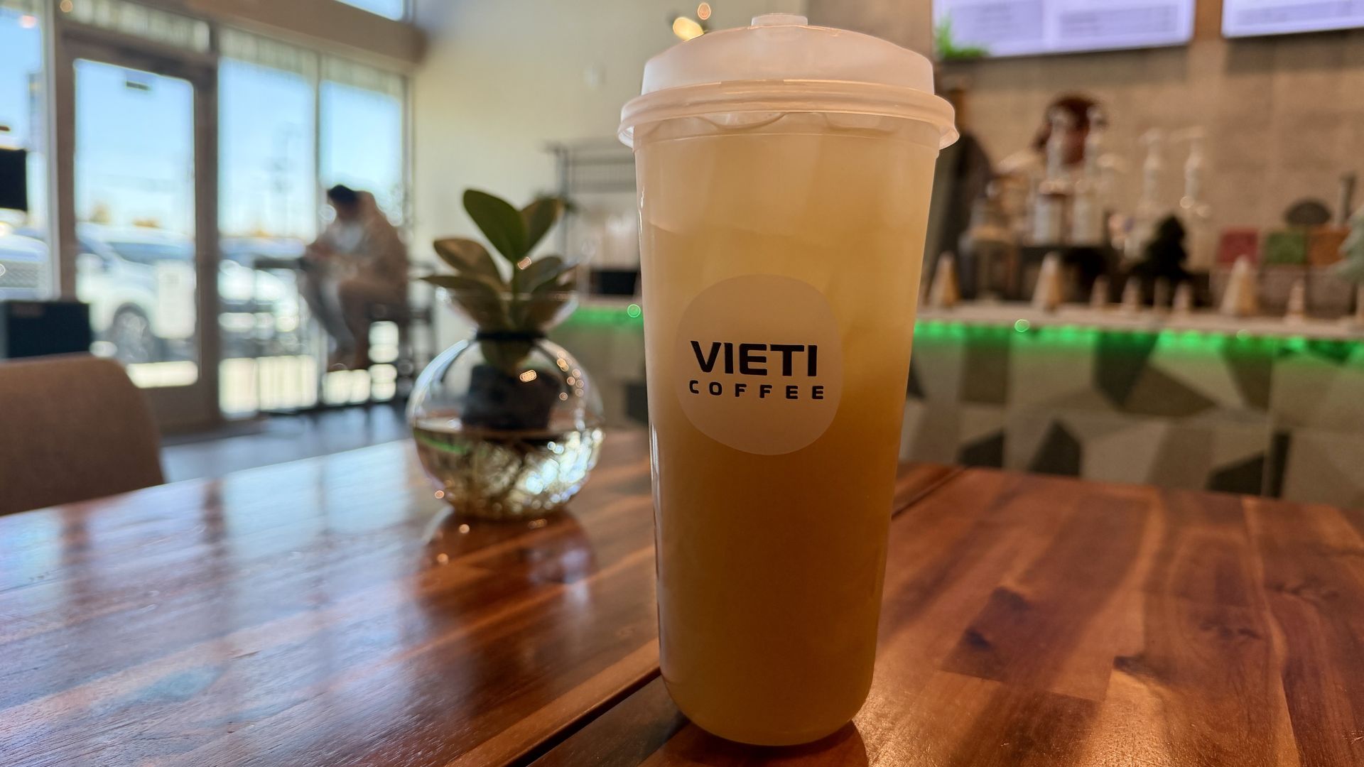 Tall iced drink cup of sugarcane juice from Vieti Coffee on a wooden table in a bright cafe, with a small glass vase plant and blurred background of a person and counter with green lighting.