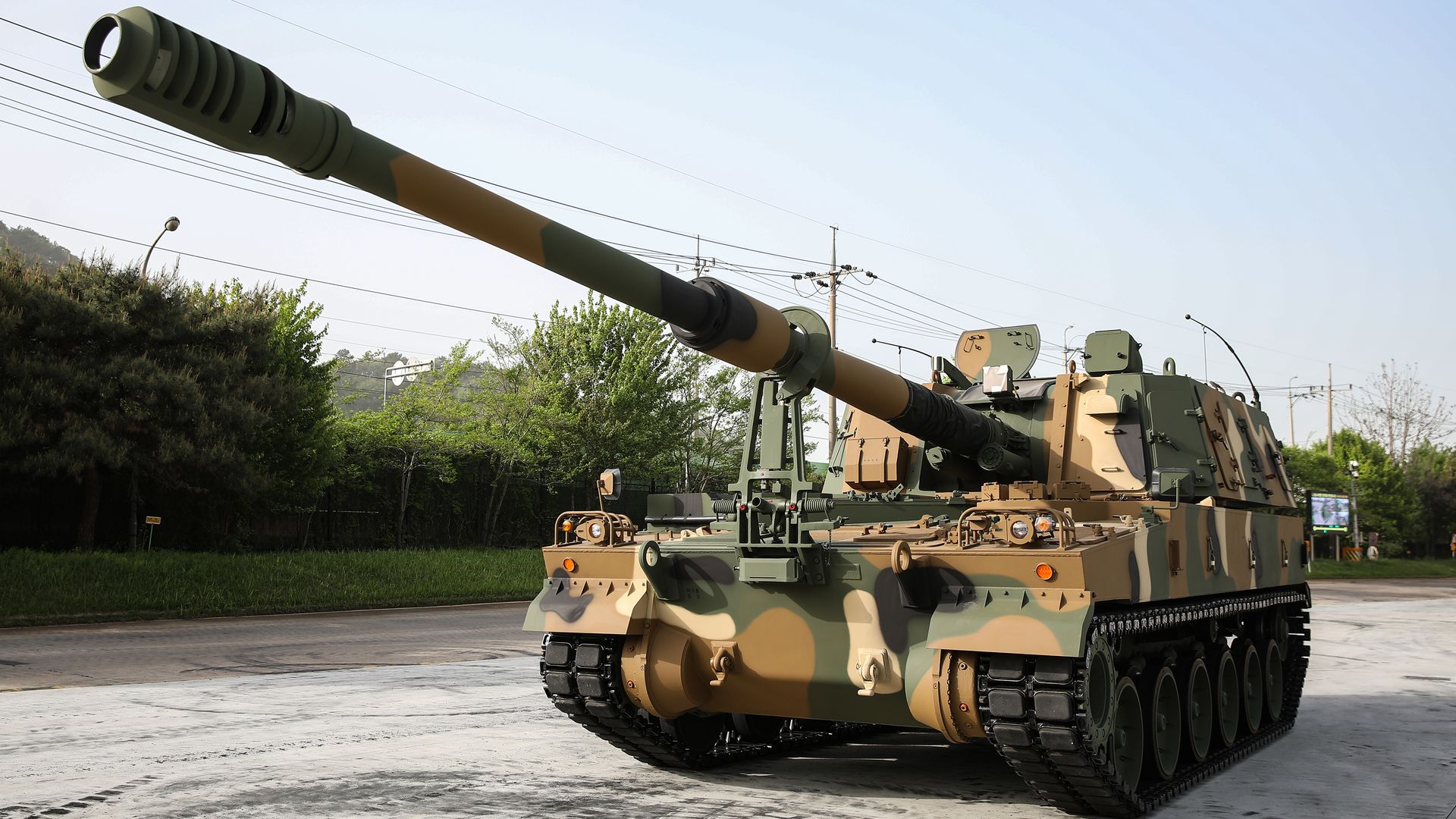 A Hanwha artillery vehicle sits in a parking lot in South Korea. A line of trees is seen behind it. The sky is blue.