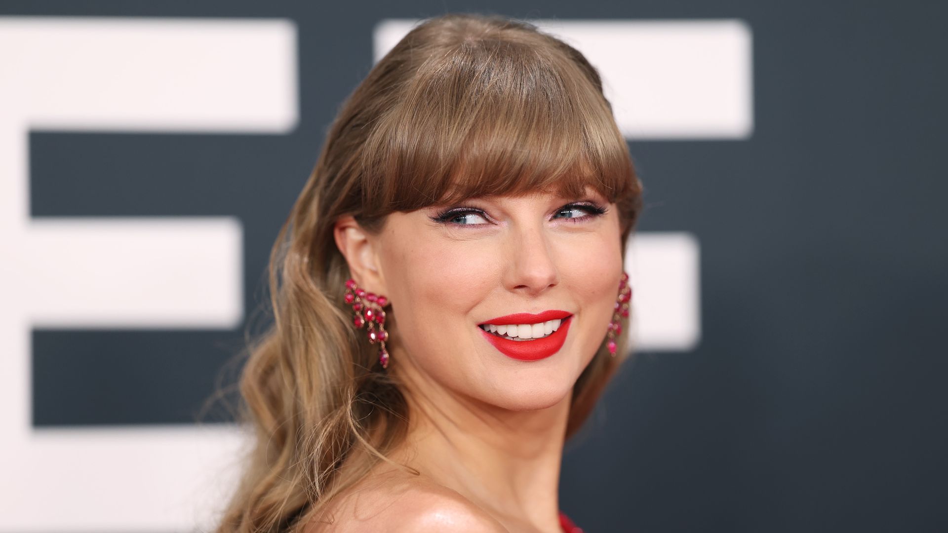 Woman with light brown wavy hair and bangs, wearing red lipstick, red earrings, and a red outfit, smiling and looking to her left against a dark background with white text.