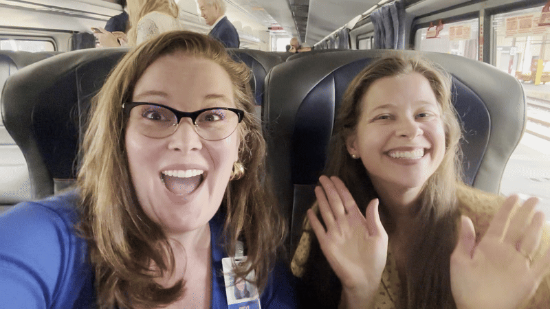 Two smiling women sitting on a train with black seats; one wearing glasses and a blue shirt, the other with long hair and a beige top, both waving at the camera.