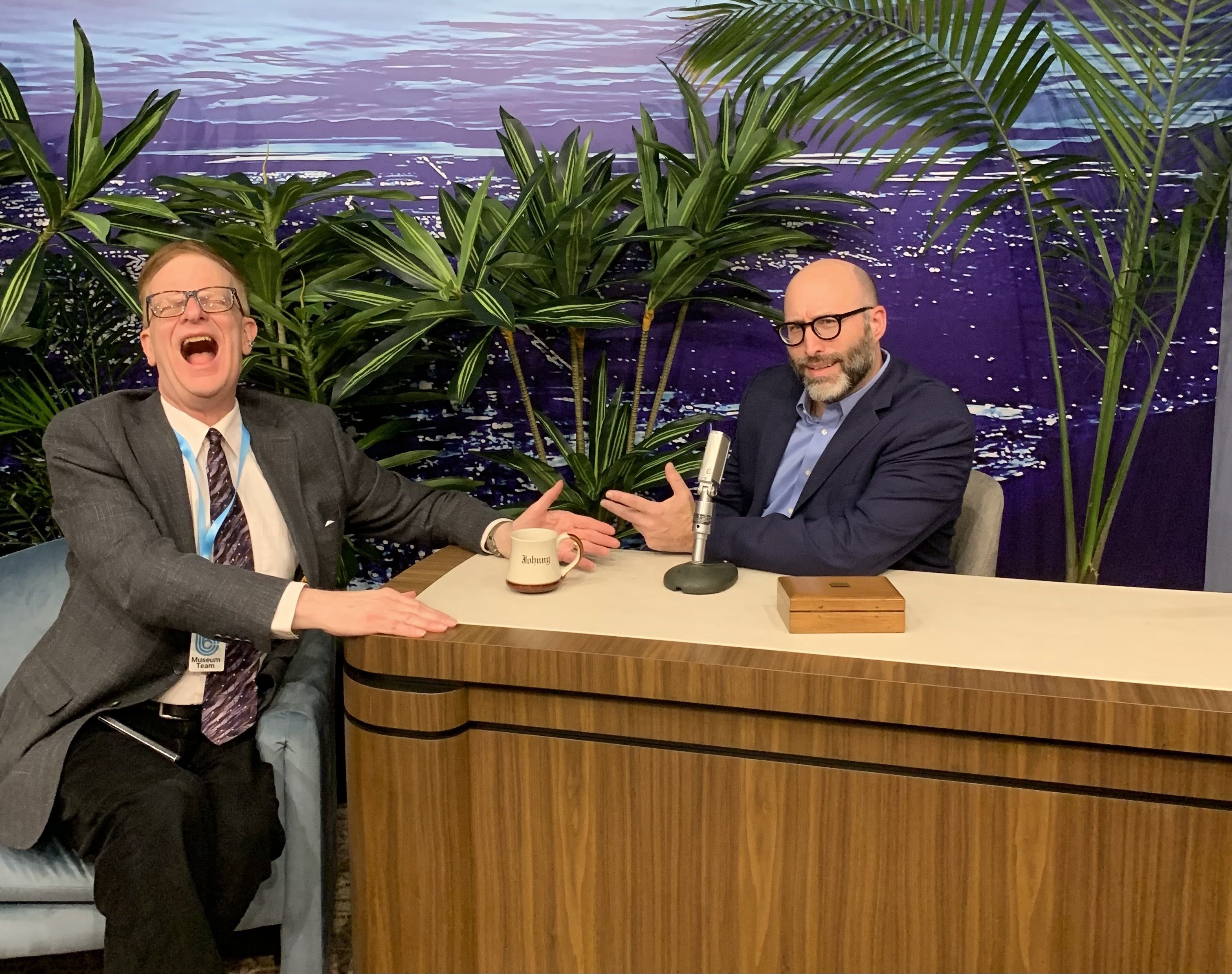 Two men in suits laughing and gesturing at a wooden talk show desk with a microphone, mug labeled "Johnny," tropical plants, and a purple ocean backdrop.