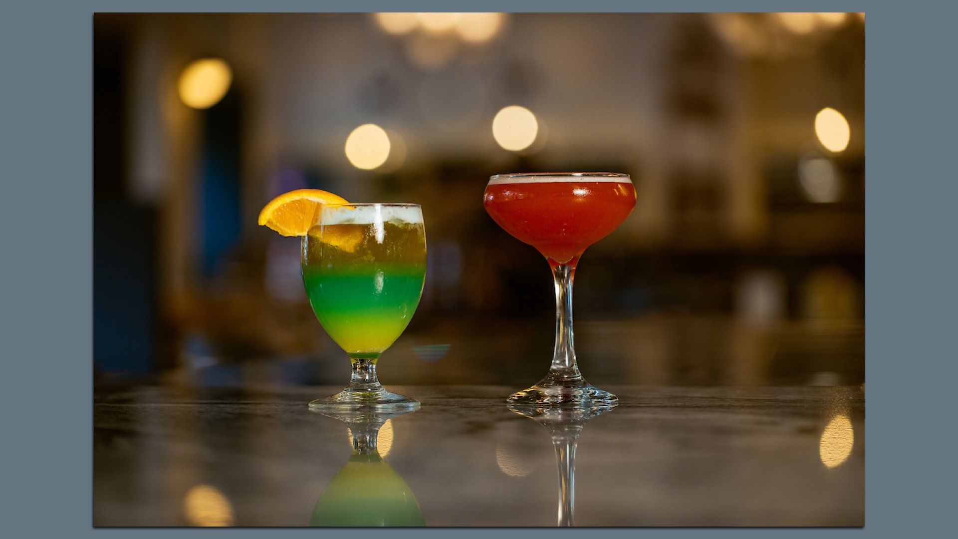 Two cocktails on a bar: a layered rainbow drink with an orange wedge on the left and a red coupe on the right, with warm bokeh lights in a blurred background.