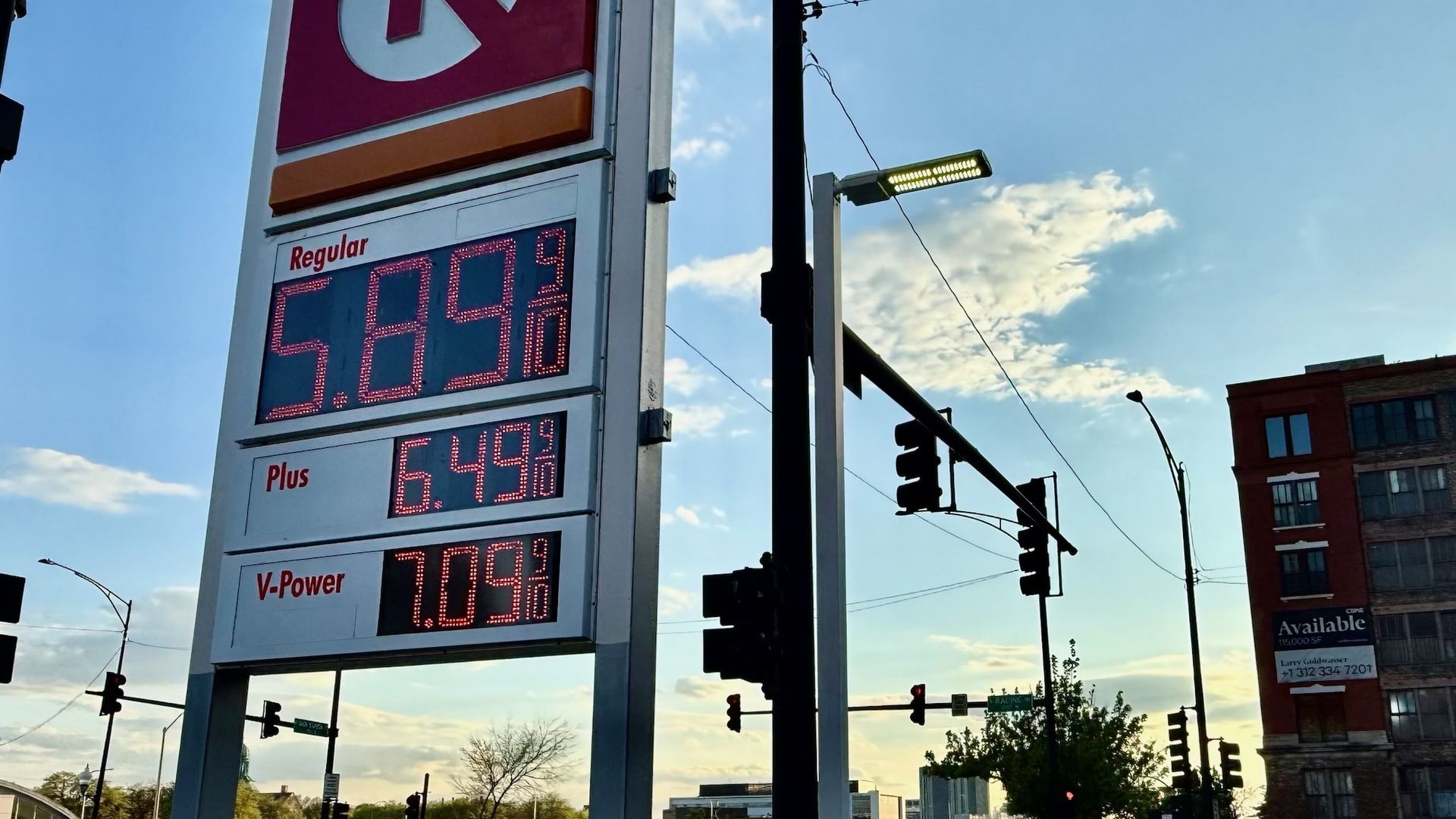 Gas station sign with a large red K logo on a pink panel. Digital red price displays show Regular, Plus, and V-Power; city street with power lines, traffic lights, and a brick building in view.