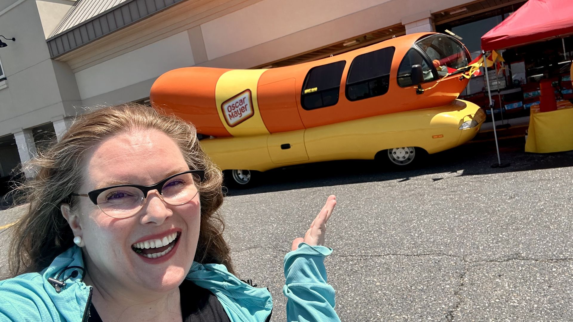Selfie of a smiling woman with glasses in a turquoise jacket, foreground, as an orange and yellow Oscar Mayer Weinermobile sits parked beside a red canopy market stall.