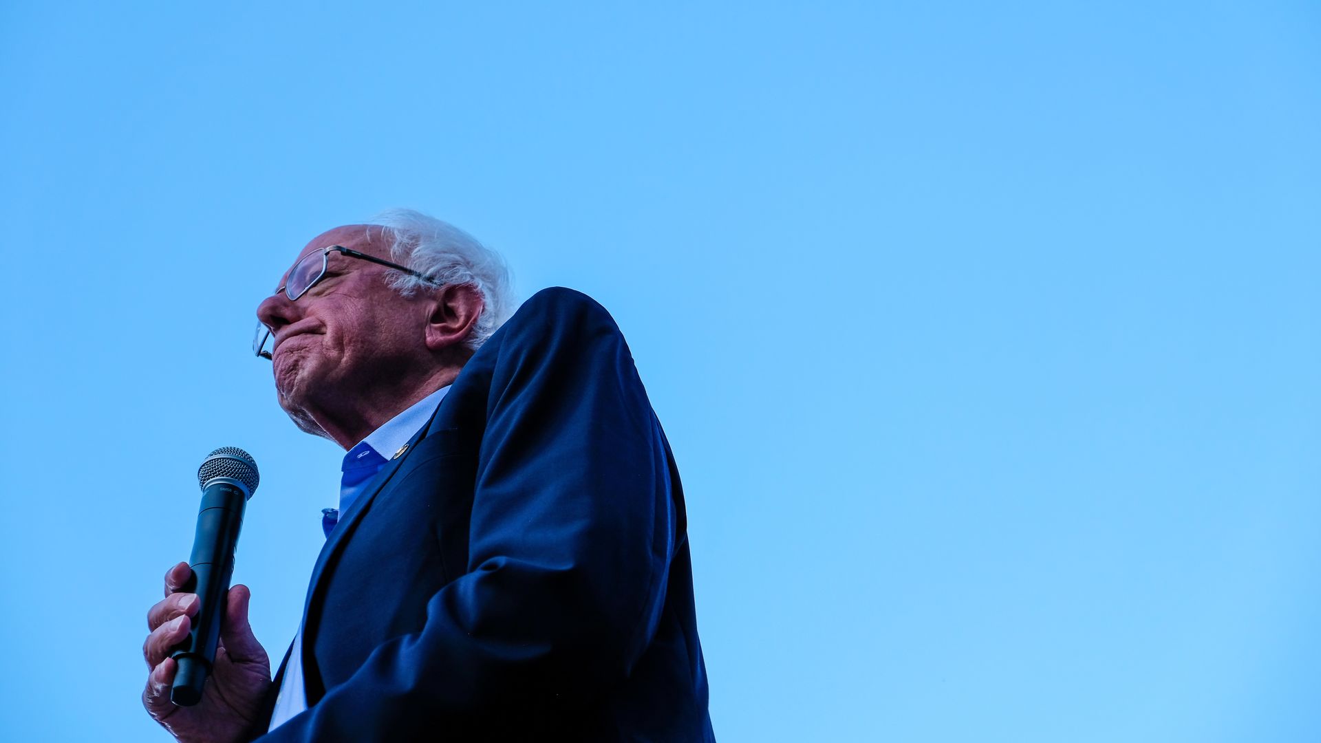 In this image, the camera looks sharply up at Bernie as he holds a microphone and stands under a clear blue sky.