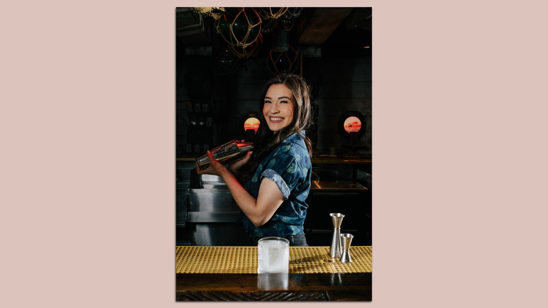 Smiling bartender in a blue floral shirt shakes a cocktail behind a dark bar; she holds a metal shaker. On the counter is an ice-filled glass with nearby bar tools.