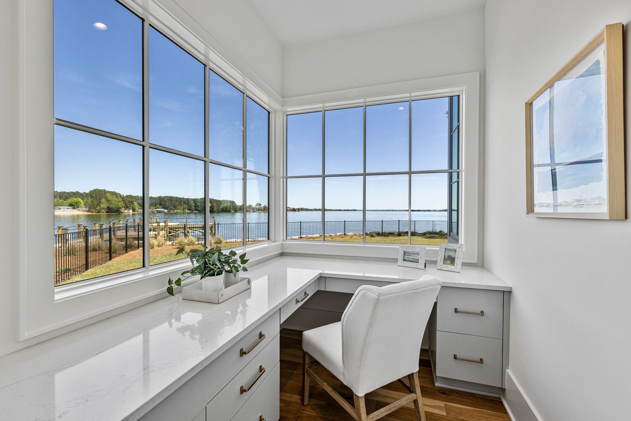 Modern white corner desk with drawers and a white chair next to large windows showing a lake, green trees, and a clear blue sky. A green plant and framed photos sit on the desk.