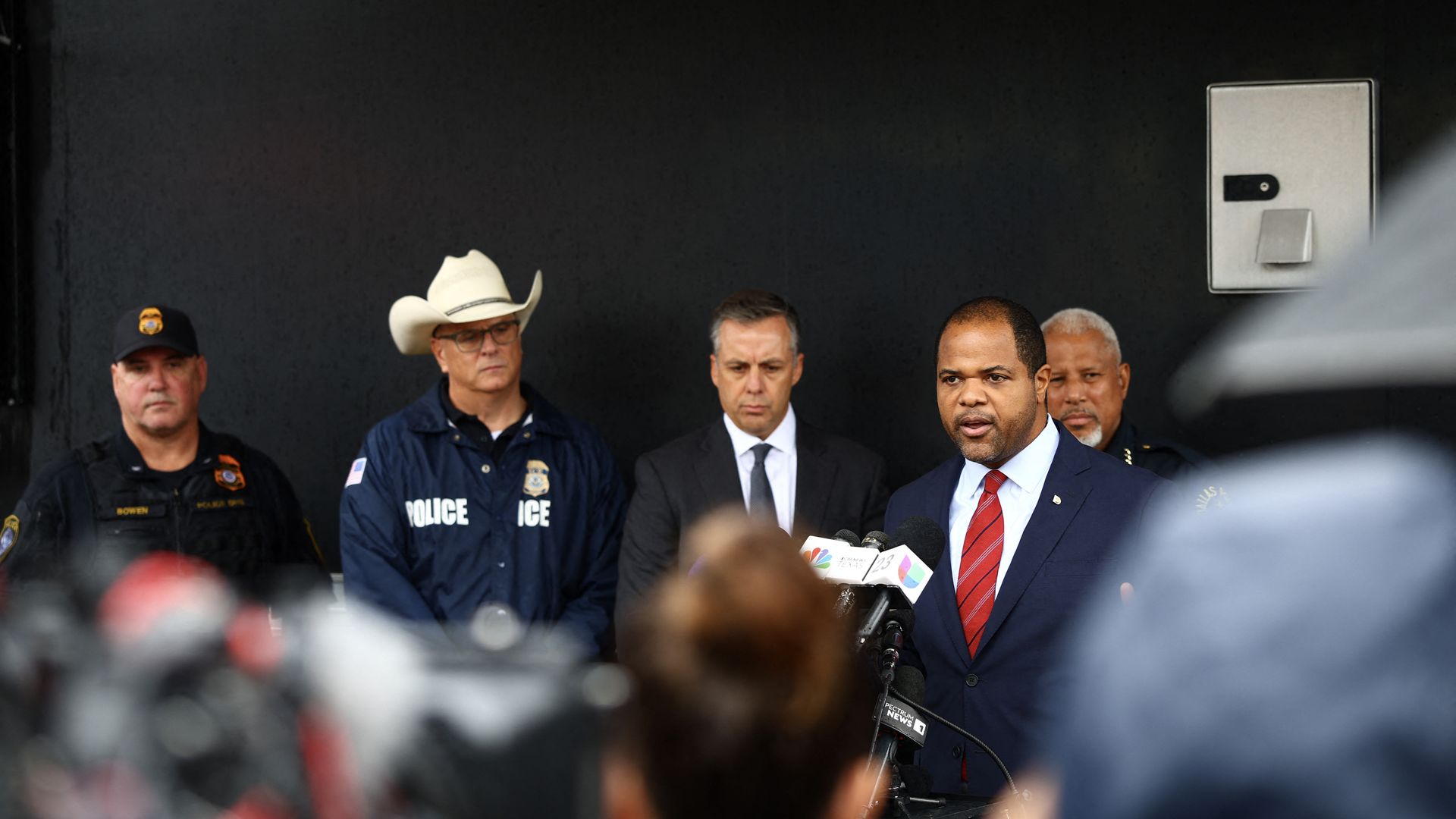 A man in a dark suit and a red tie speaks at a news conference while four other men in suits and law enforcement uniforms stand behind him.