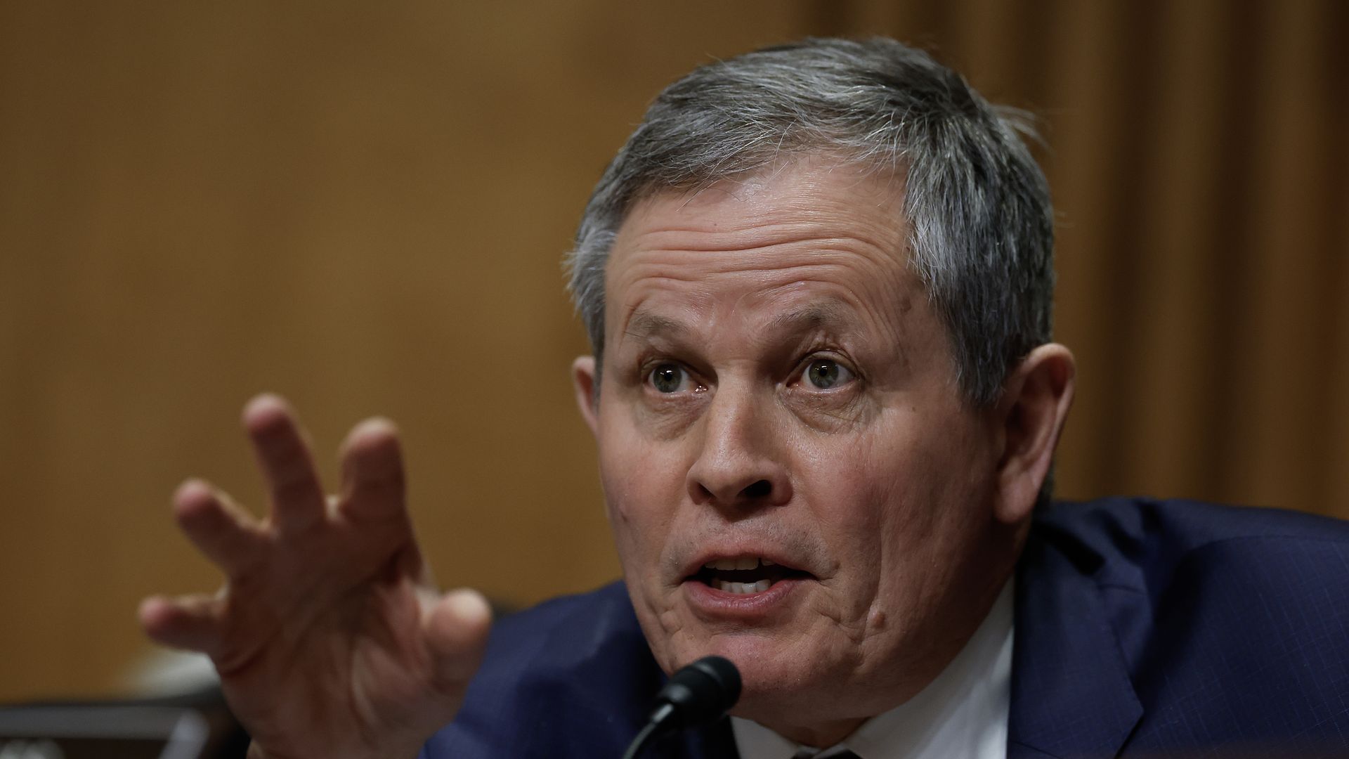 Sen. Steve Daines, with gray hair wearing a dark suit and red striped tie speaking into a microphone, gesturing with his hand, brown background.