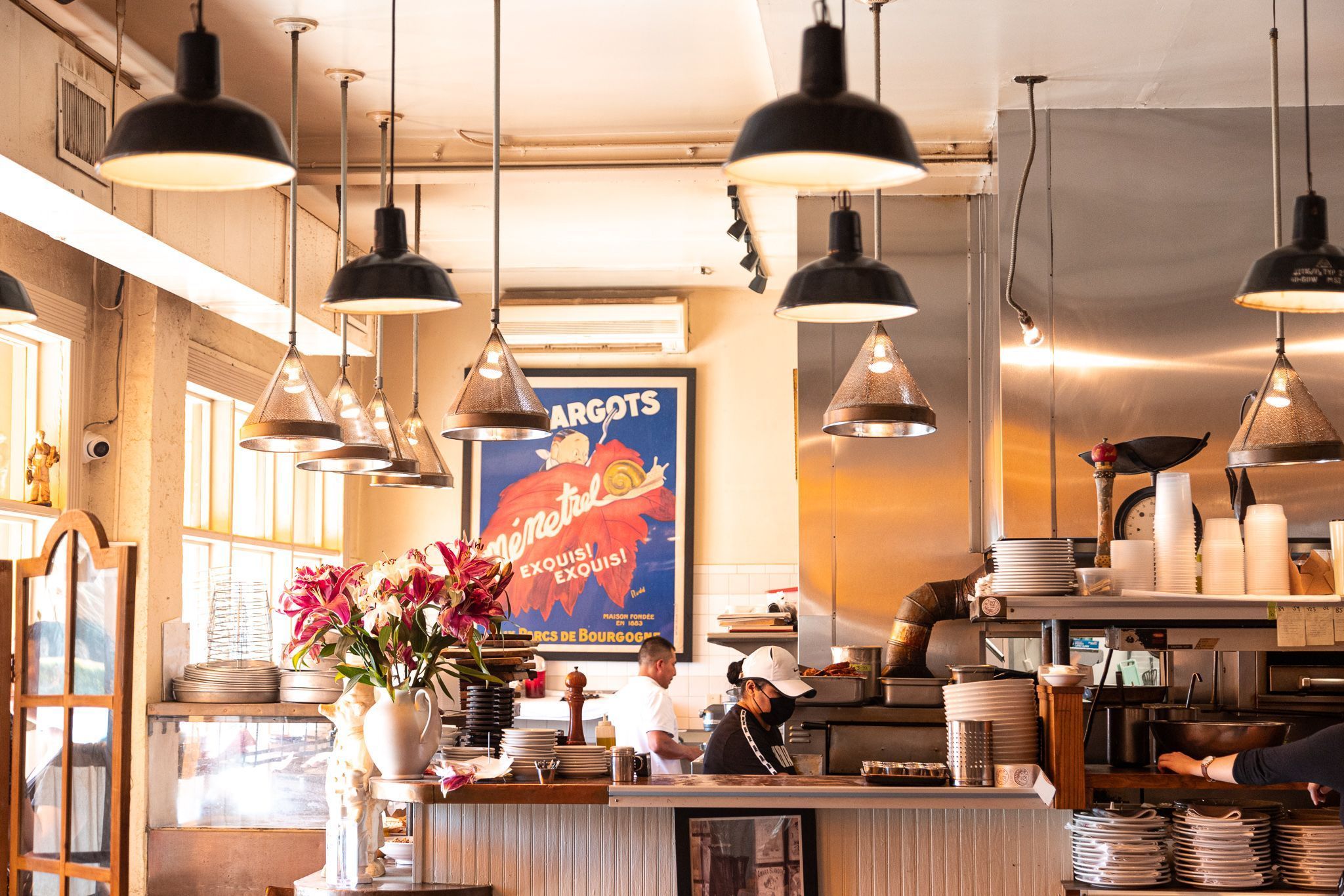 Interior of a bright cafe with black industrial pendant lights, white walls, and a busy open kitchen. A vase of pink lilies sits on the counter among stacked plates; staff in white and black uniforms work near stainless steel equipment.