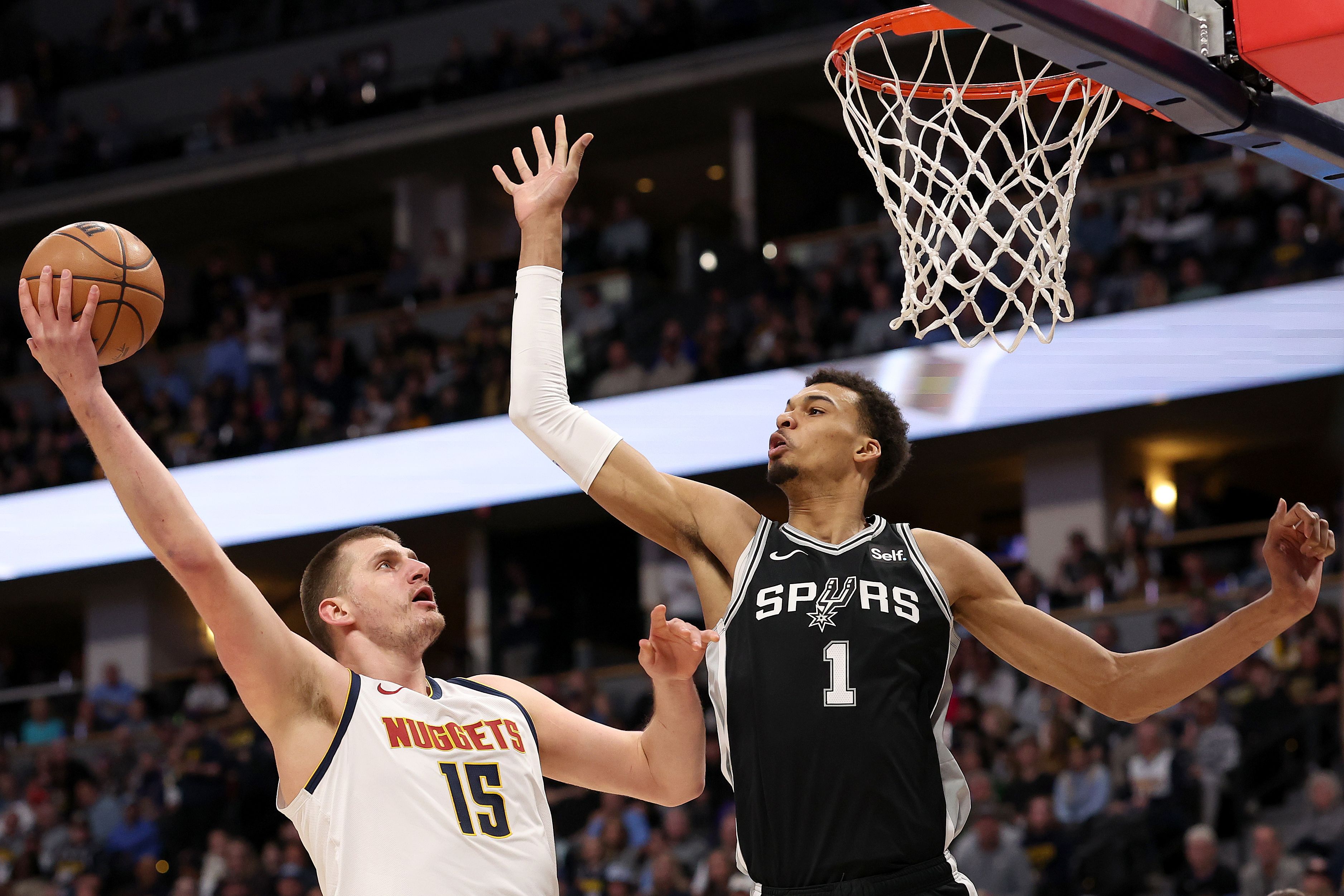 A man in a white jersey tries to shot an orange basketball over a man wearing a black jersey with the word SPURS on it. Both players are near a basket with white laces.