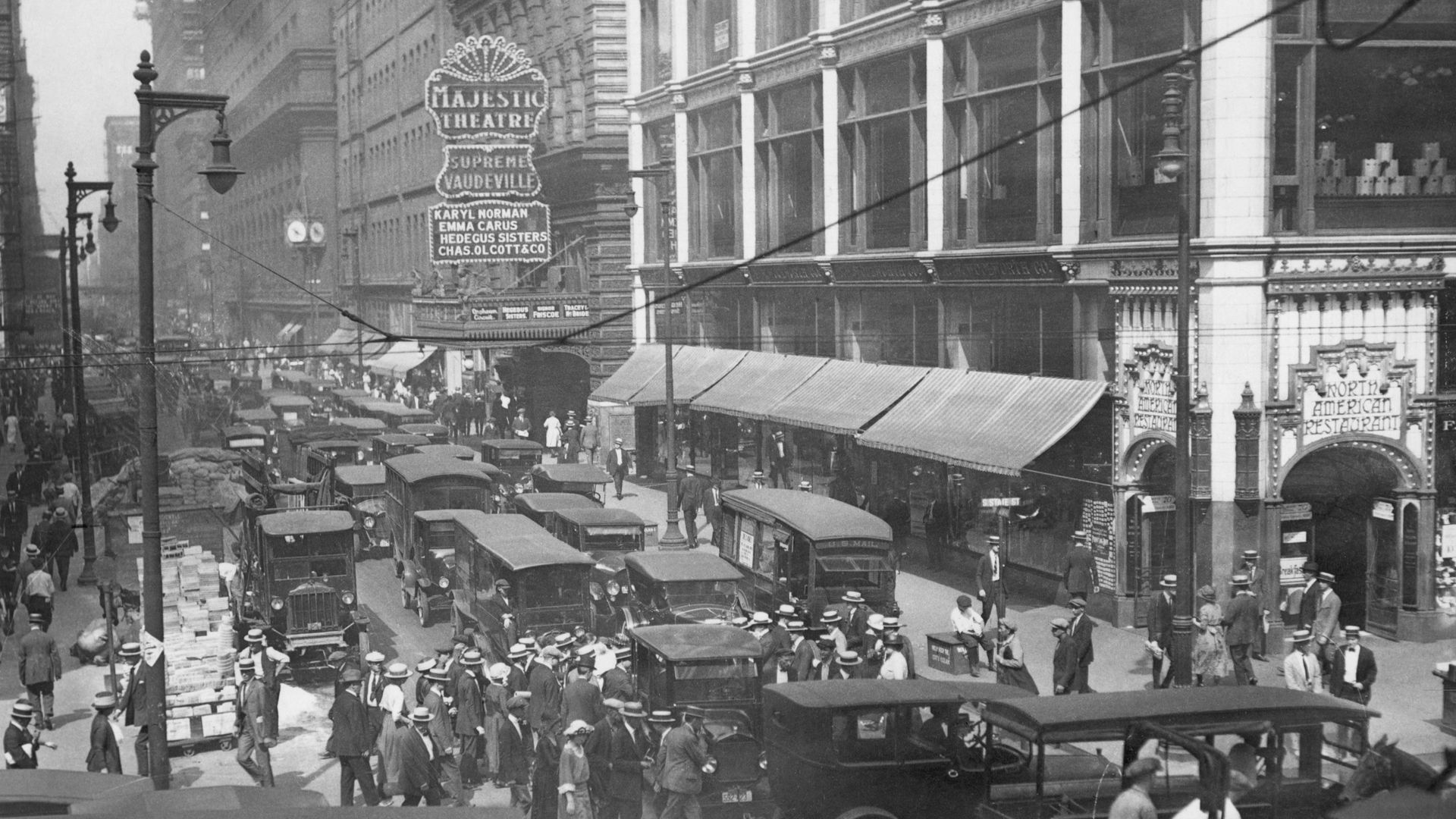 Black-and-white early-20th-century street scene with a crowded avenue, vintage cars and pedestrians, and a marquee reading "Majestic Theatre" on a grand ornate building.