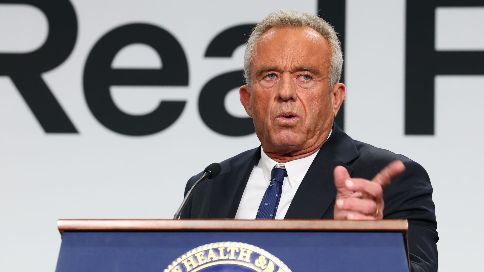 A gray-haired Secretary of Health and Human Services Robert F. Kennedy Jr., in a dark suit and navy tie, speaks and gestures at a podium with a Department of Health & Human Services emblem, against a white background with large black text.