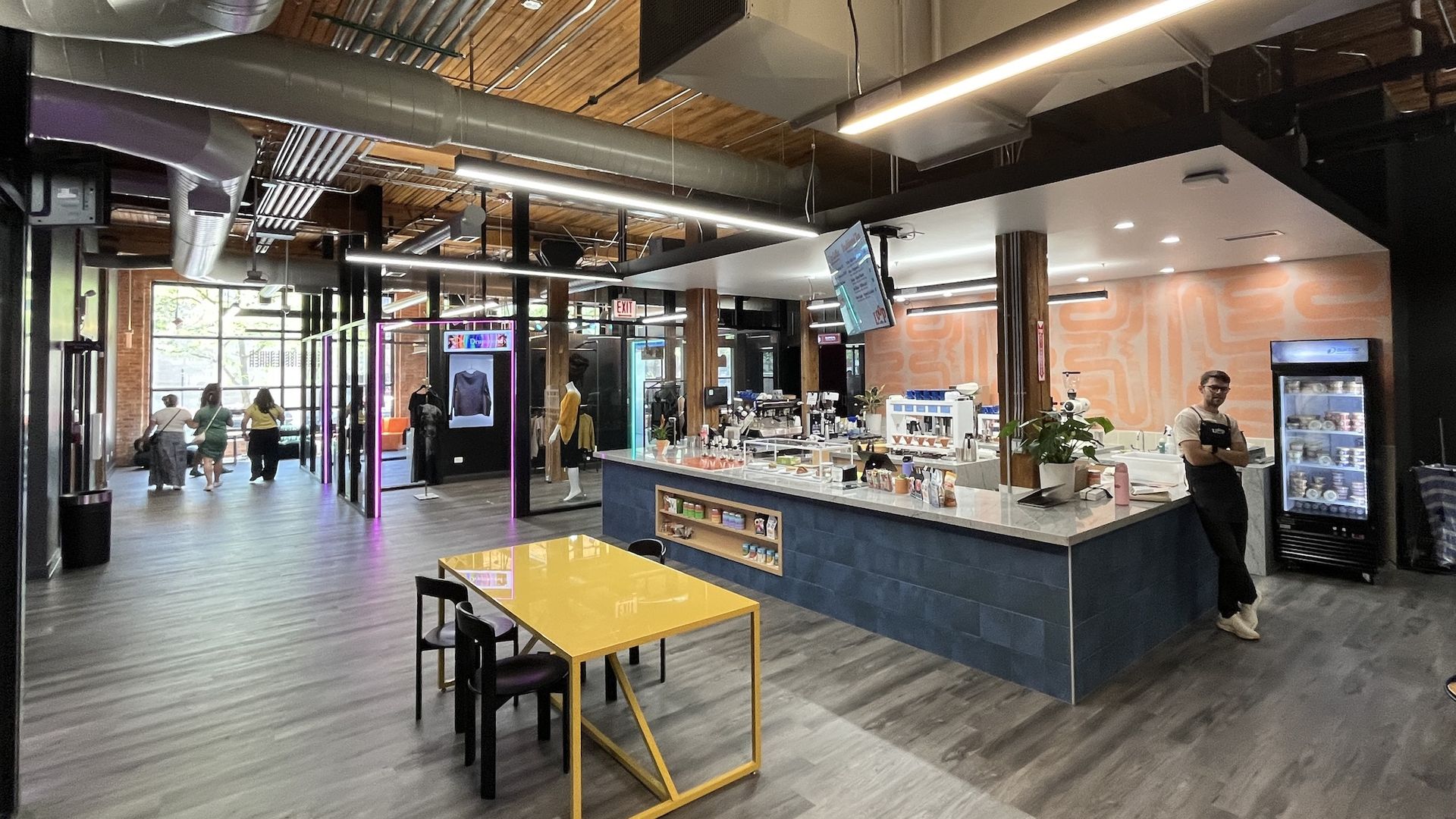 Industrial-style indoor space with exposed ducts, wooden ceiling, and bright linear lights. A counter with a blue base and glass display, a yellow table in the foreground, and people walking toward glass doors.