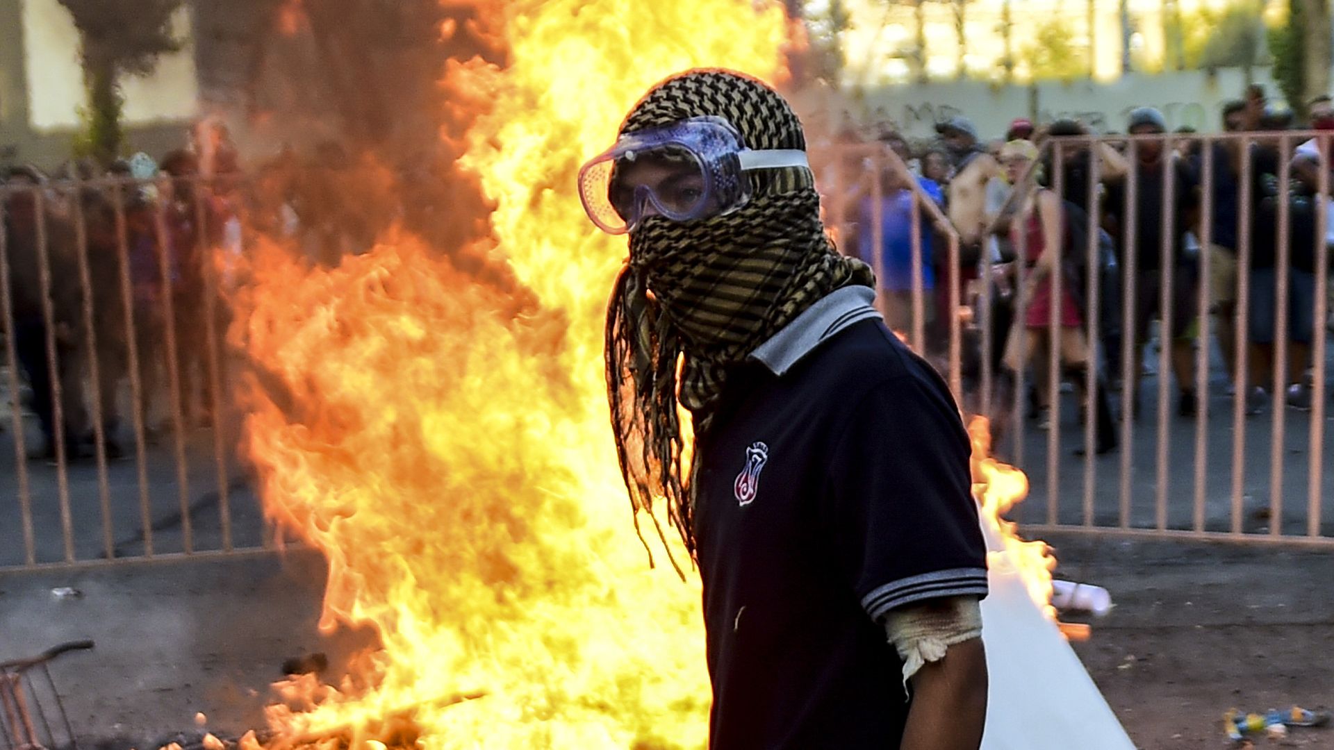 In this image, a young man stands in front of a large fire while wearing goggles and a head scarf