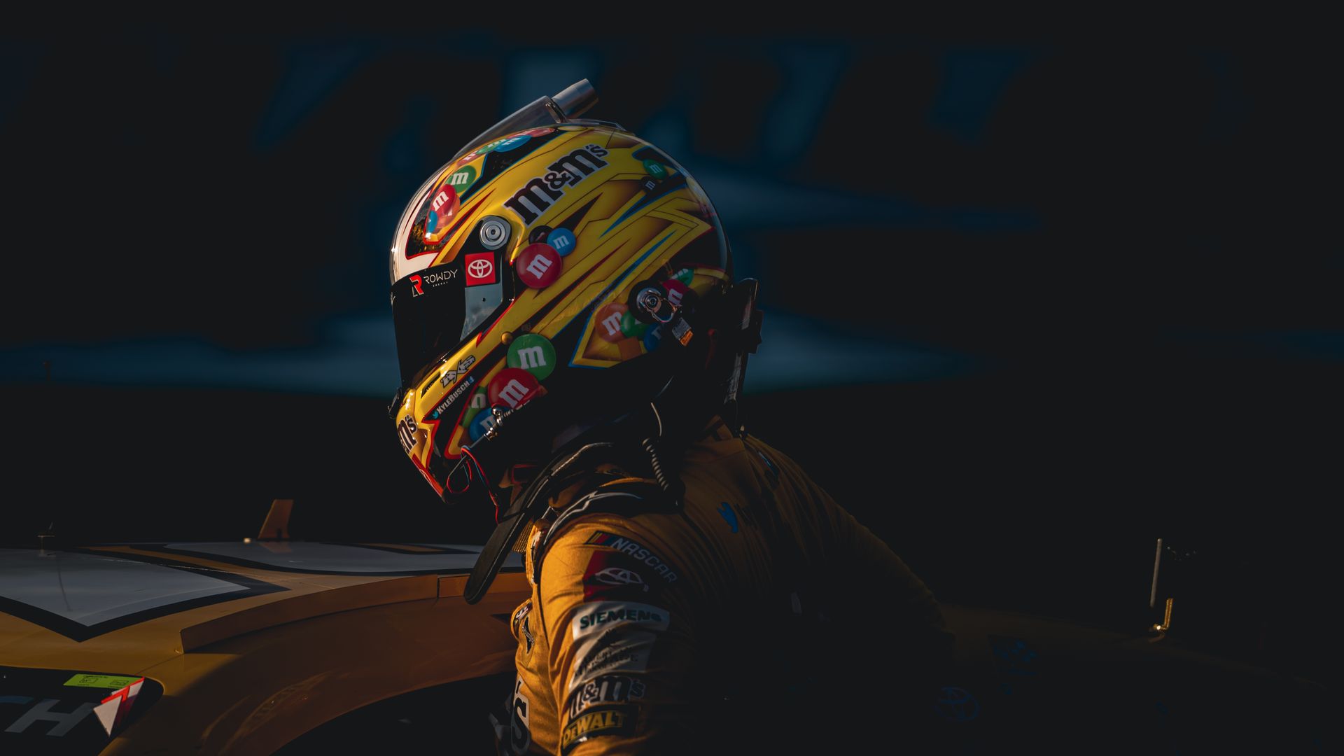 Close-up of a yellow racing helmet covered in colorful M&M's stickers and sponsor logos, worn by a driver in a matching suit against a dark background.