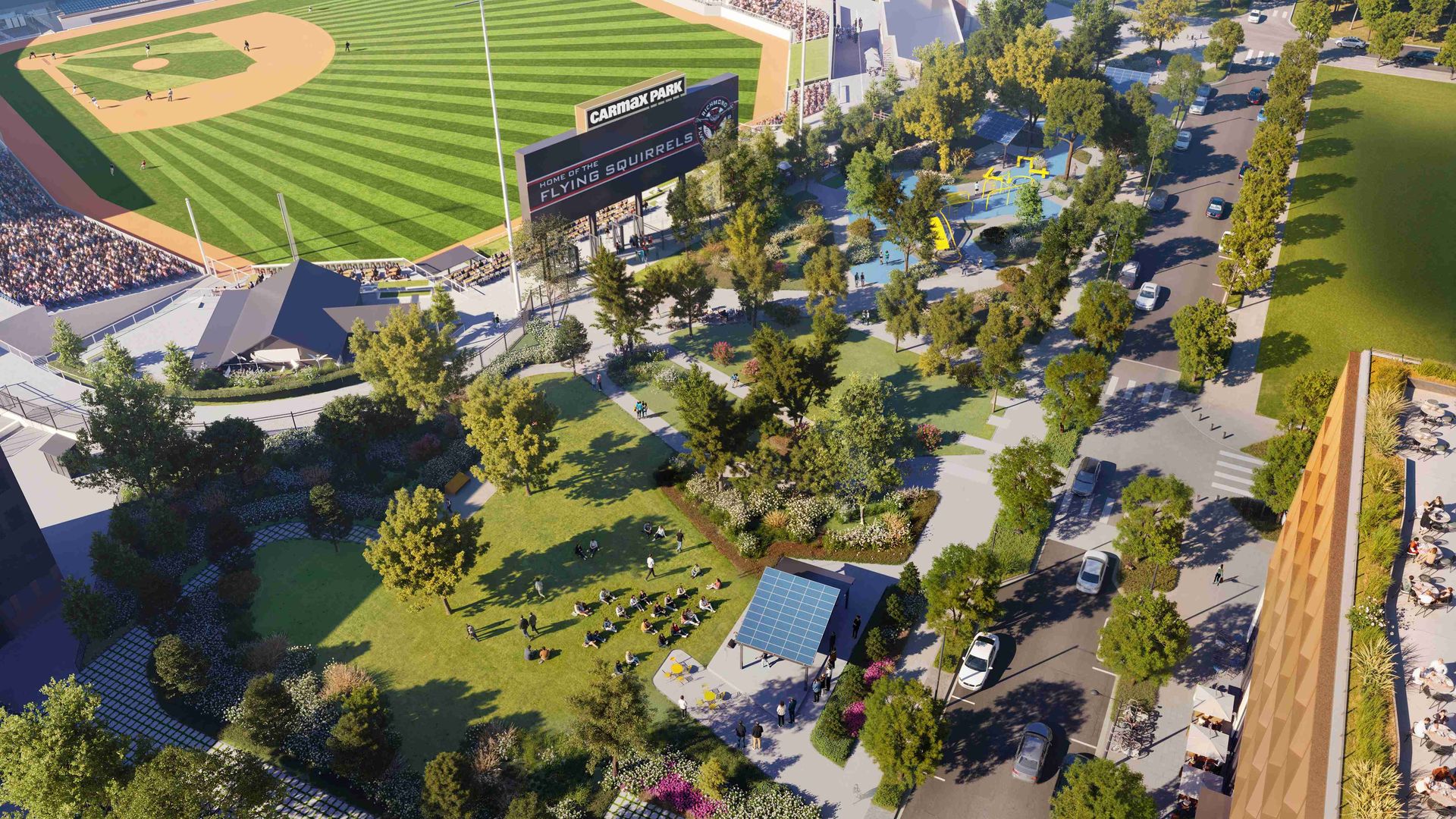 Aerial view of CarMax Park baseball stadium with a striped green field and a large scoreboard reading "Home of the Flying Squirrels," surrounded by trees, a playground, and a street with cars.