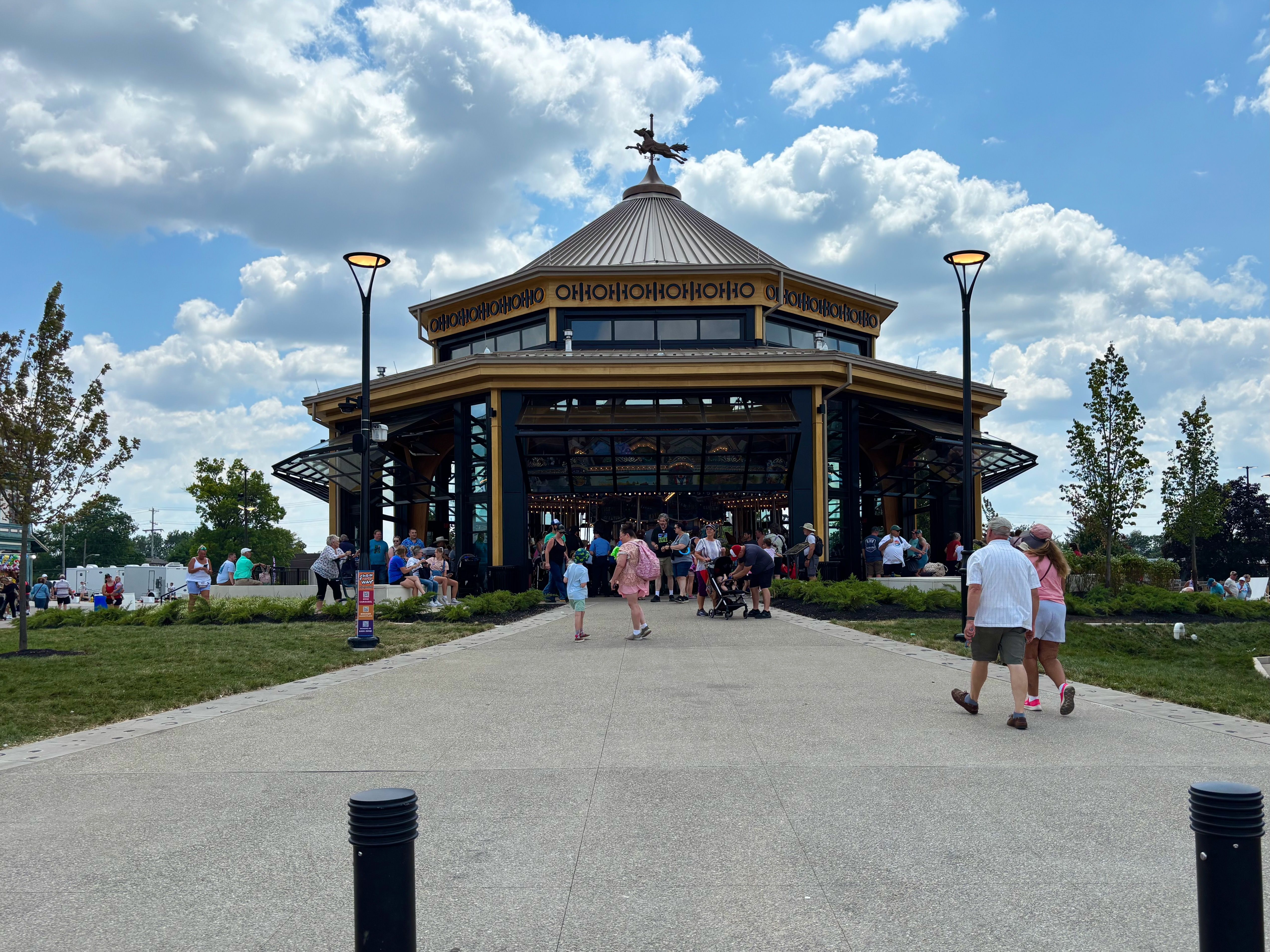 Crowds gather outside a glass-front carousel pavilion with a tall conical metal roof and statue on top, beneath a blue sky with white clouds.