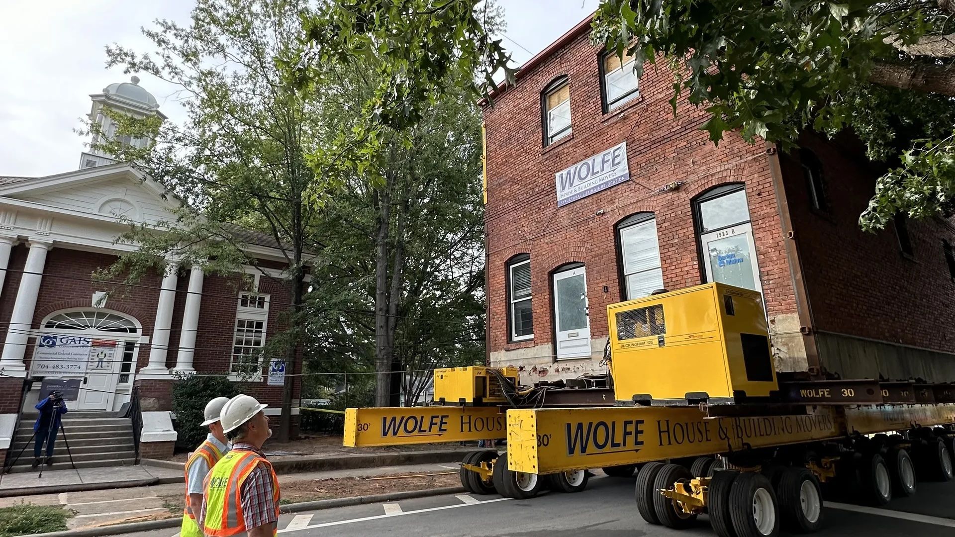 Two workers in safety vests and helmets watch a brick building being moved on a large yellow platform labeled "WOLFE" outdoors near a red brick building with white columns.