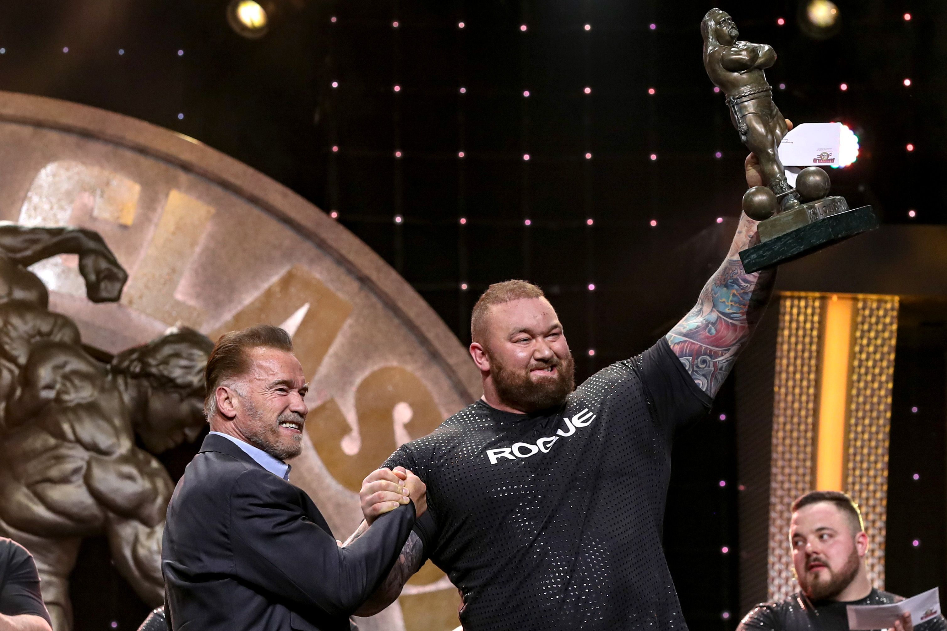 Arnold Schwarzenegger presents a trophy to the Arnold Strongman Classic winner, who holds it in the air