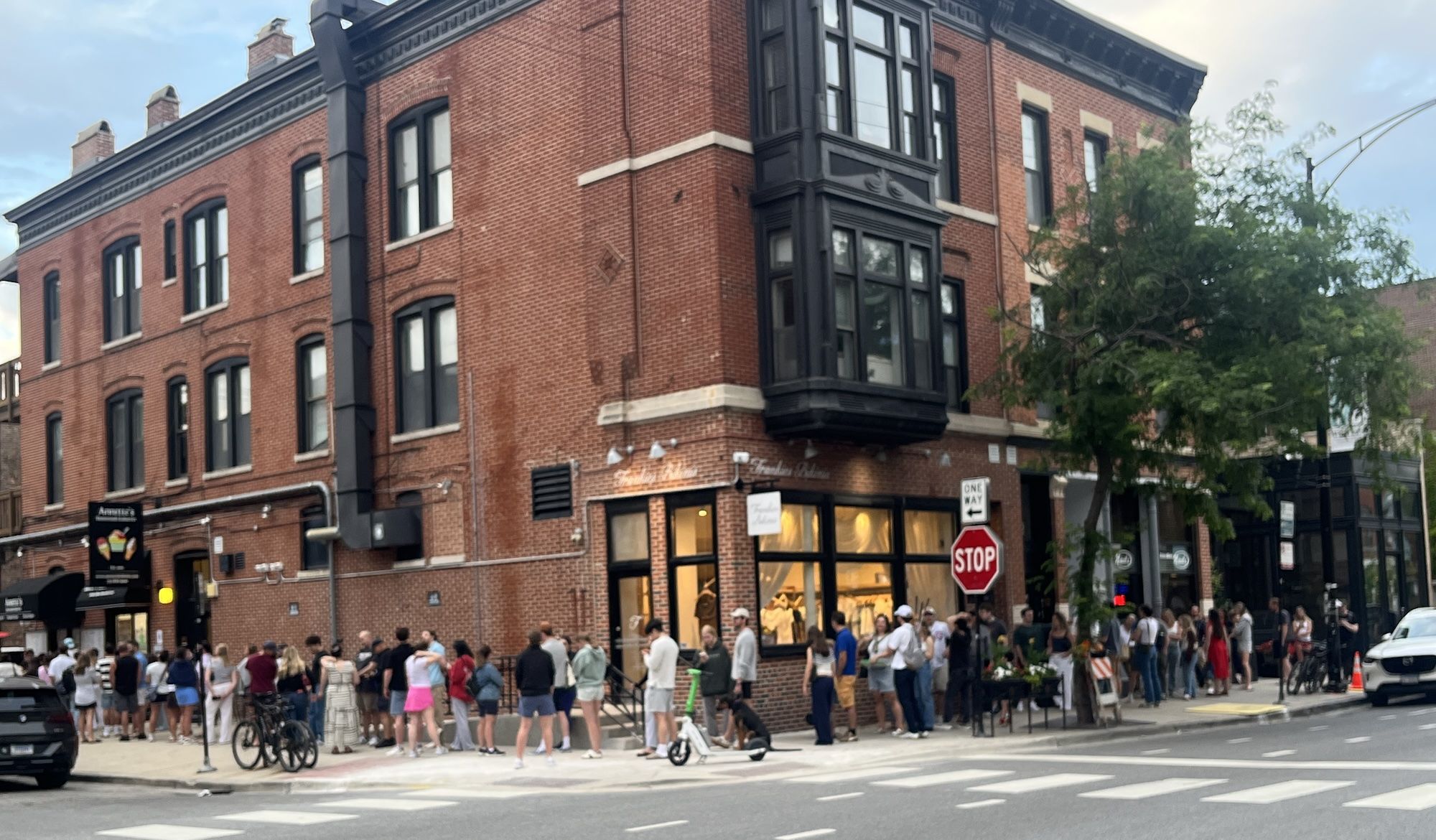 Red brick corner building with black bay windows and storefronts; crowded sidewalk with pedestrians and bicycles, a stop sign, street signs, trees, and a pale blue sky above.