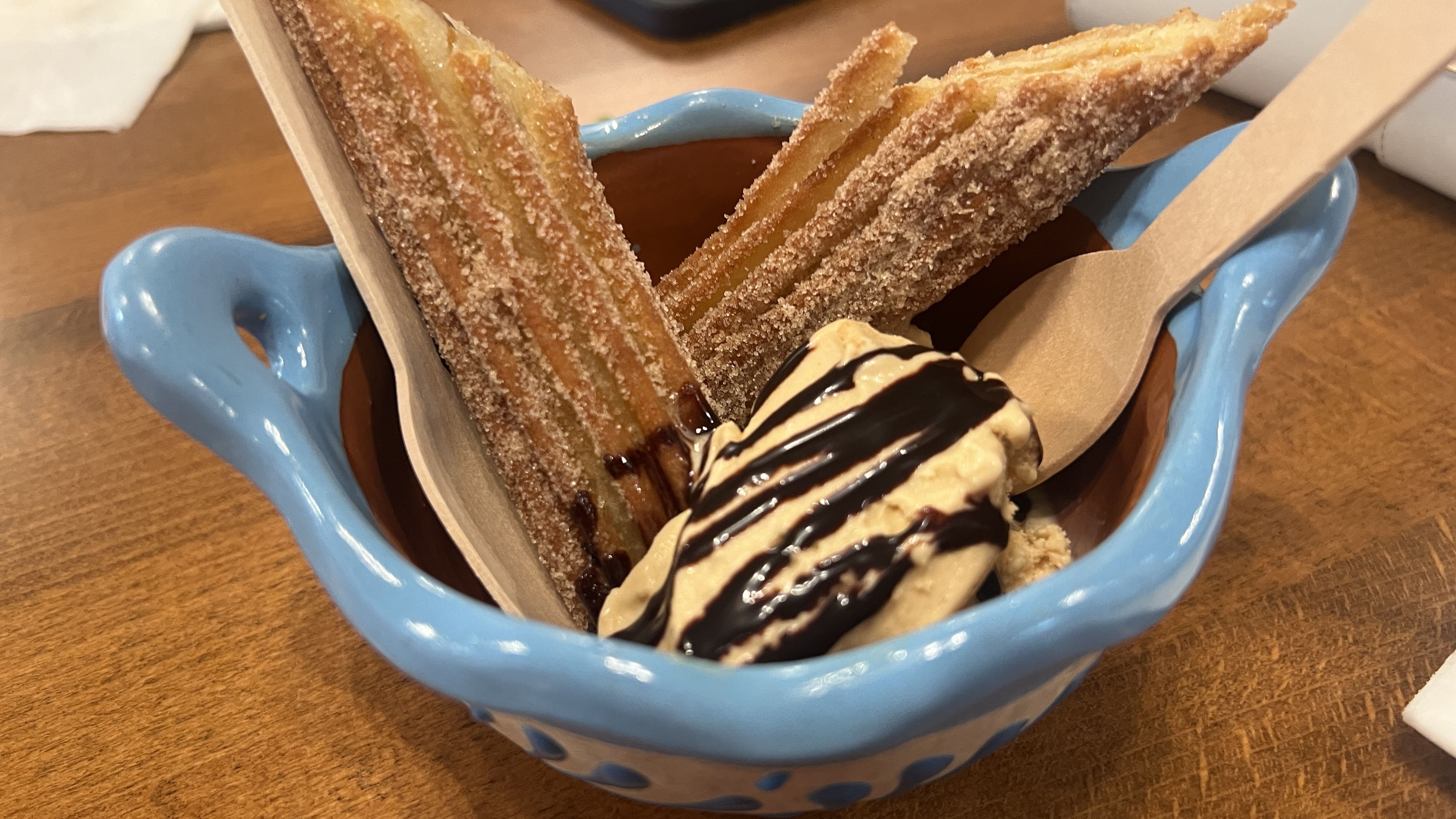 A blue bowl containing a churro broken in two, and vanilla ice cream drizzled with chocolate sauce, with wooden spoon