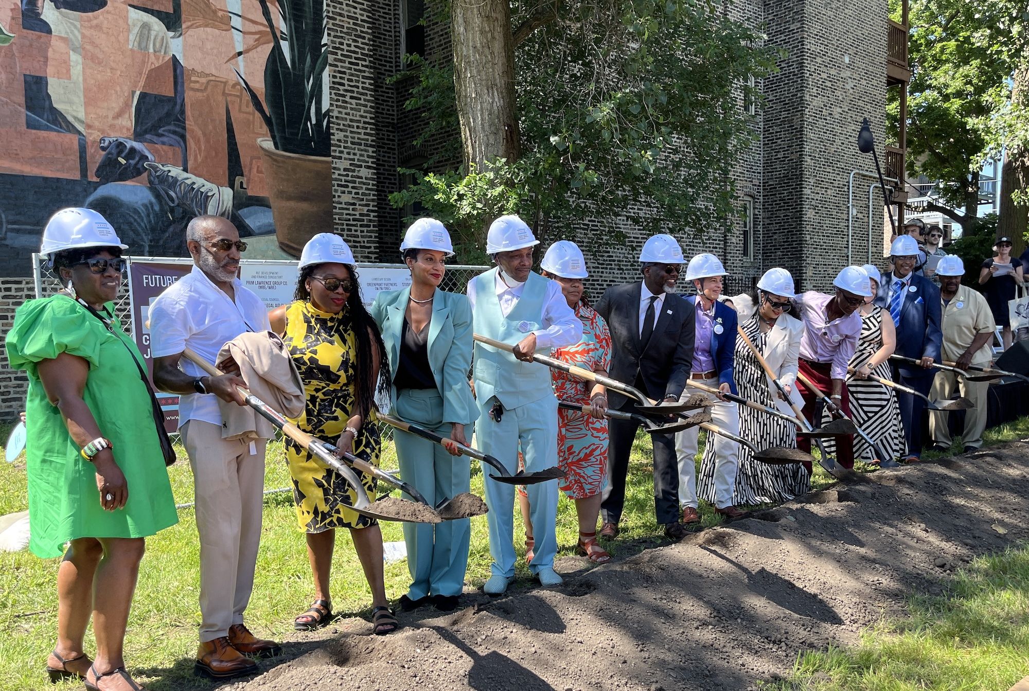 Group of diverse people in white hard hats and bright outfits line up with shovels for a groundbreaking ceremony in front of a mural and brick building, with trees nearby.