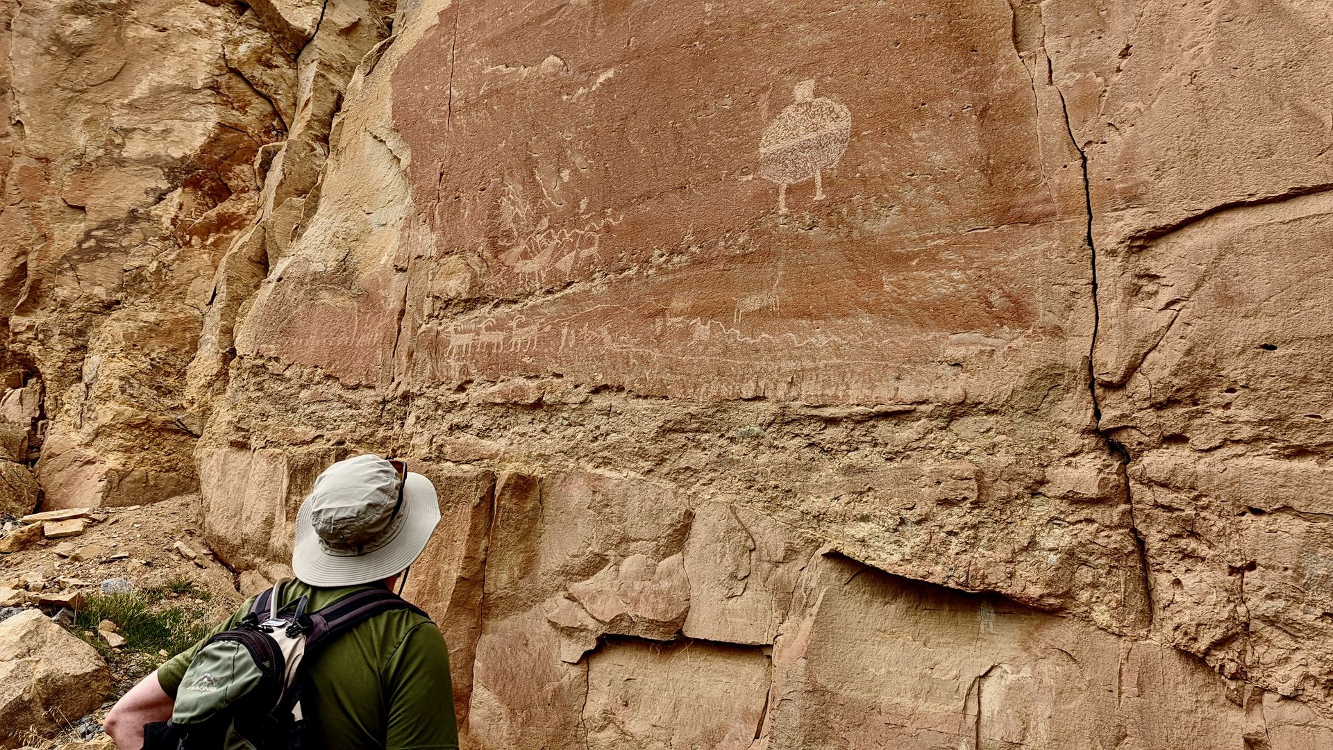 A man in a tan brimmed hat looks at rock art carved in orange stone.