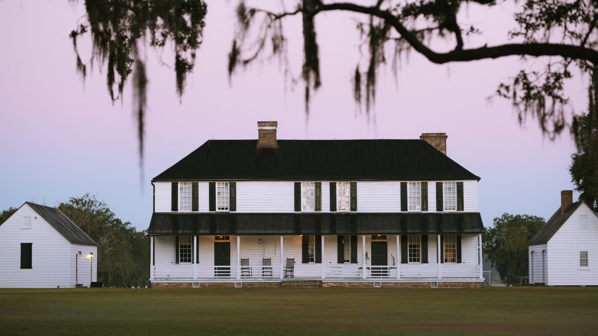 A historic white plantation-style house with black shutters and twin chimneys stands at sunset with moss-draped trees nearby.