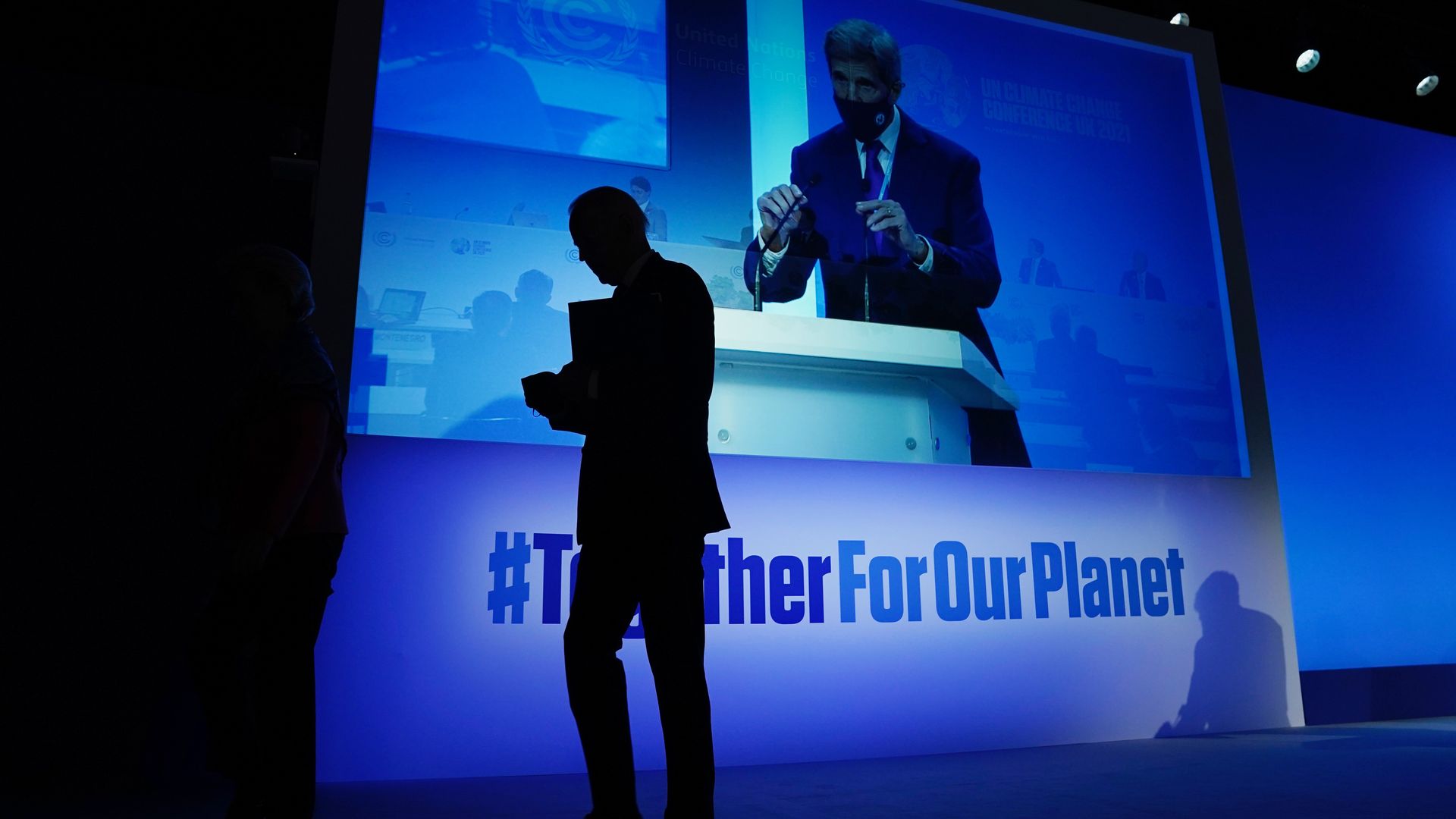 President Biden is seen in silhouette leaving the stage in Glasgow, Scotland, as special climate enjoy John Kerry steps up to the lectern.