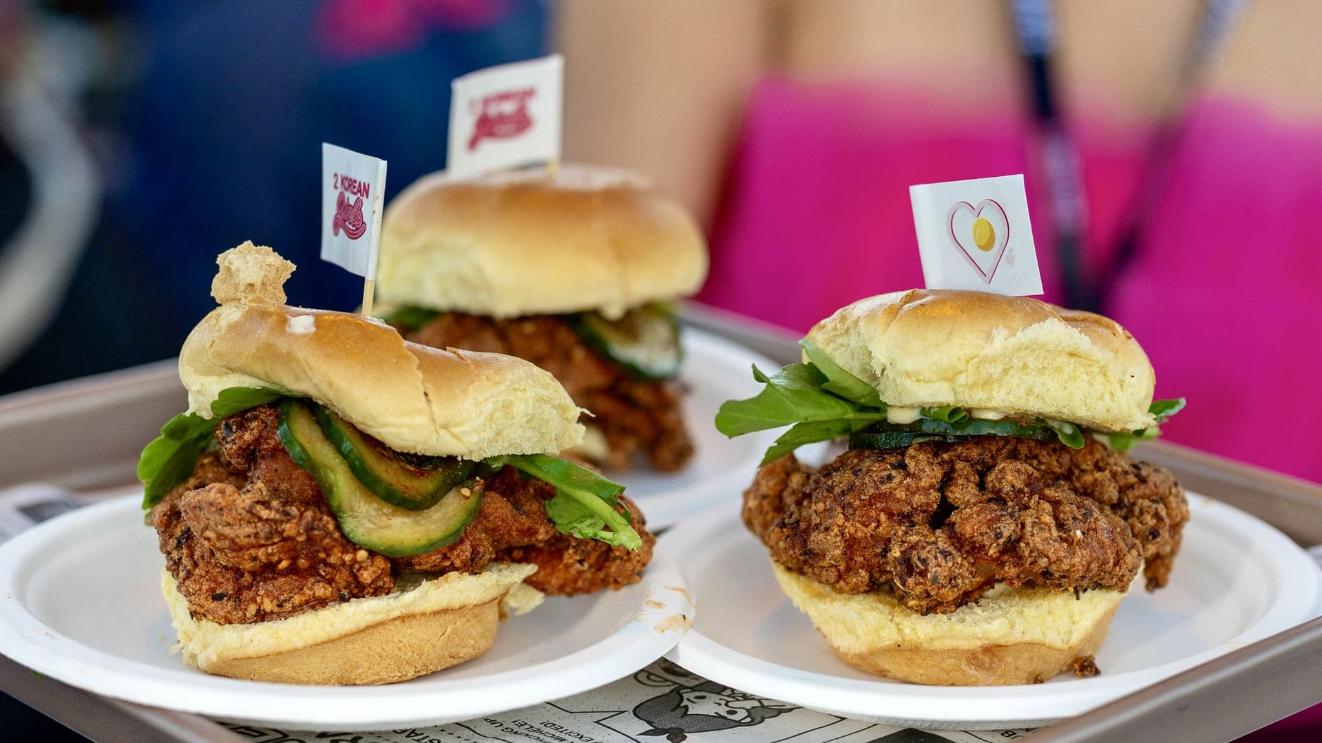 Three fried chicken sandwiches with pickles and greens on white plates, each topped with a small flag, served on a tray with newspaper-style lining.
