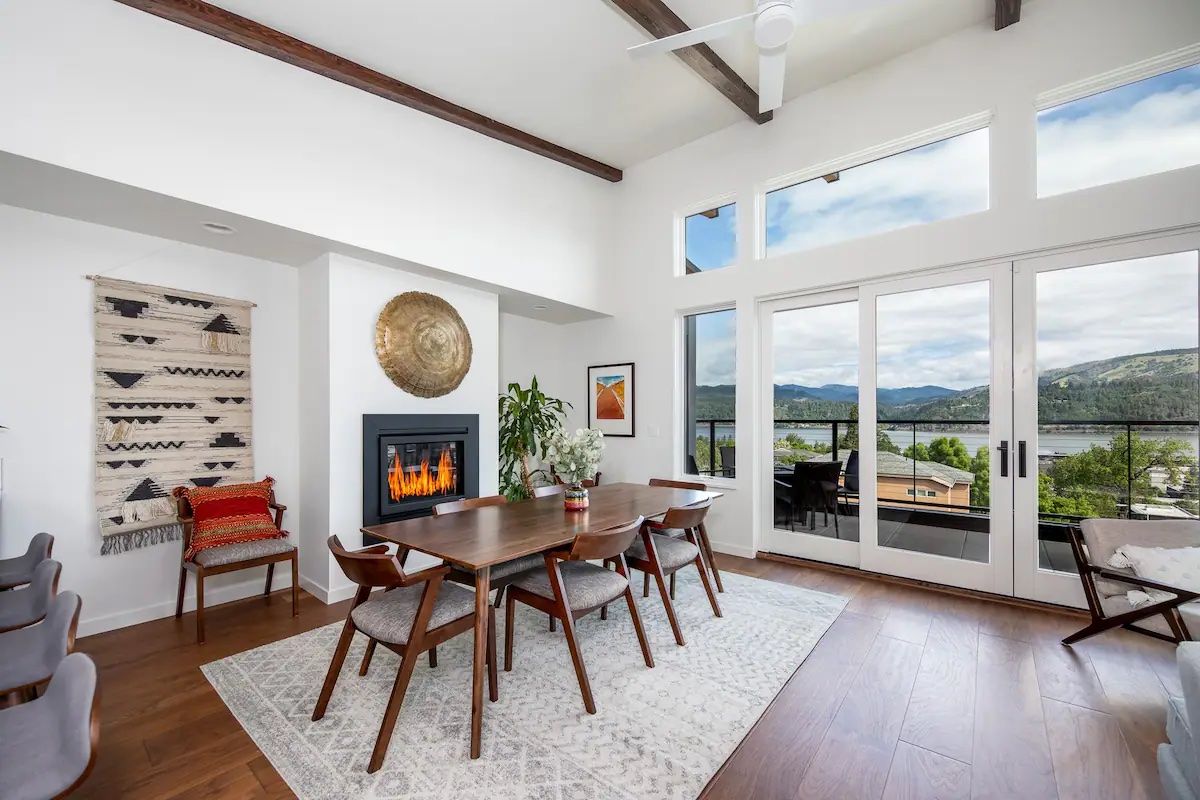 A living room with a fireplace, floor-to-ceiling windows looking out to the Columbia River, and a dining table.
